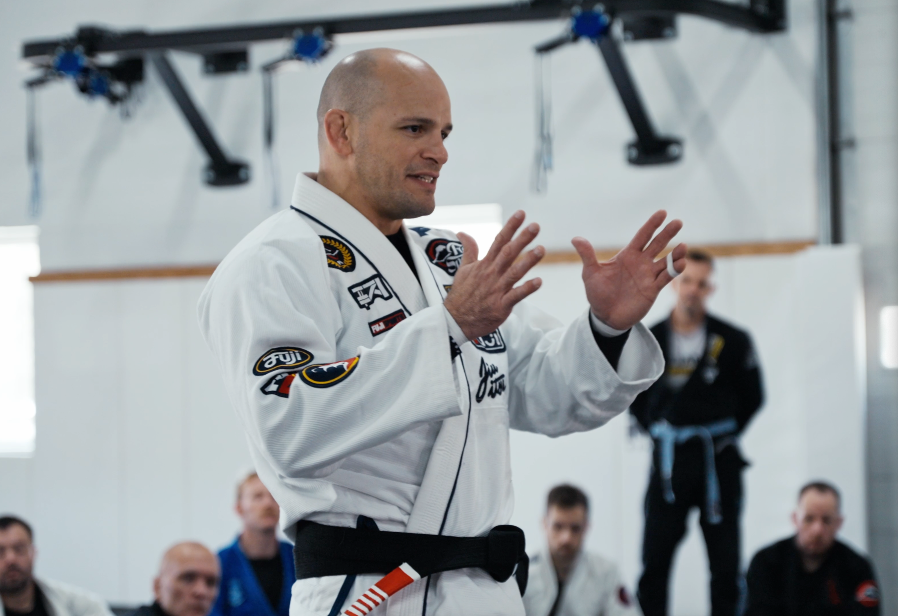 A person in a white Brazilian jiu-jitsu gi with a black belt speaks and gestures with both hands during a martial arts class, with attentive students seated in the background.