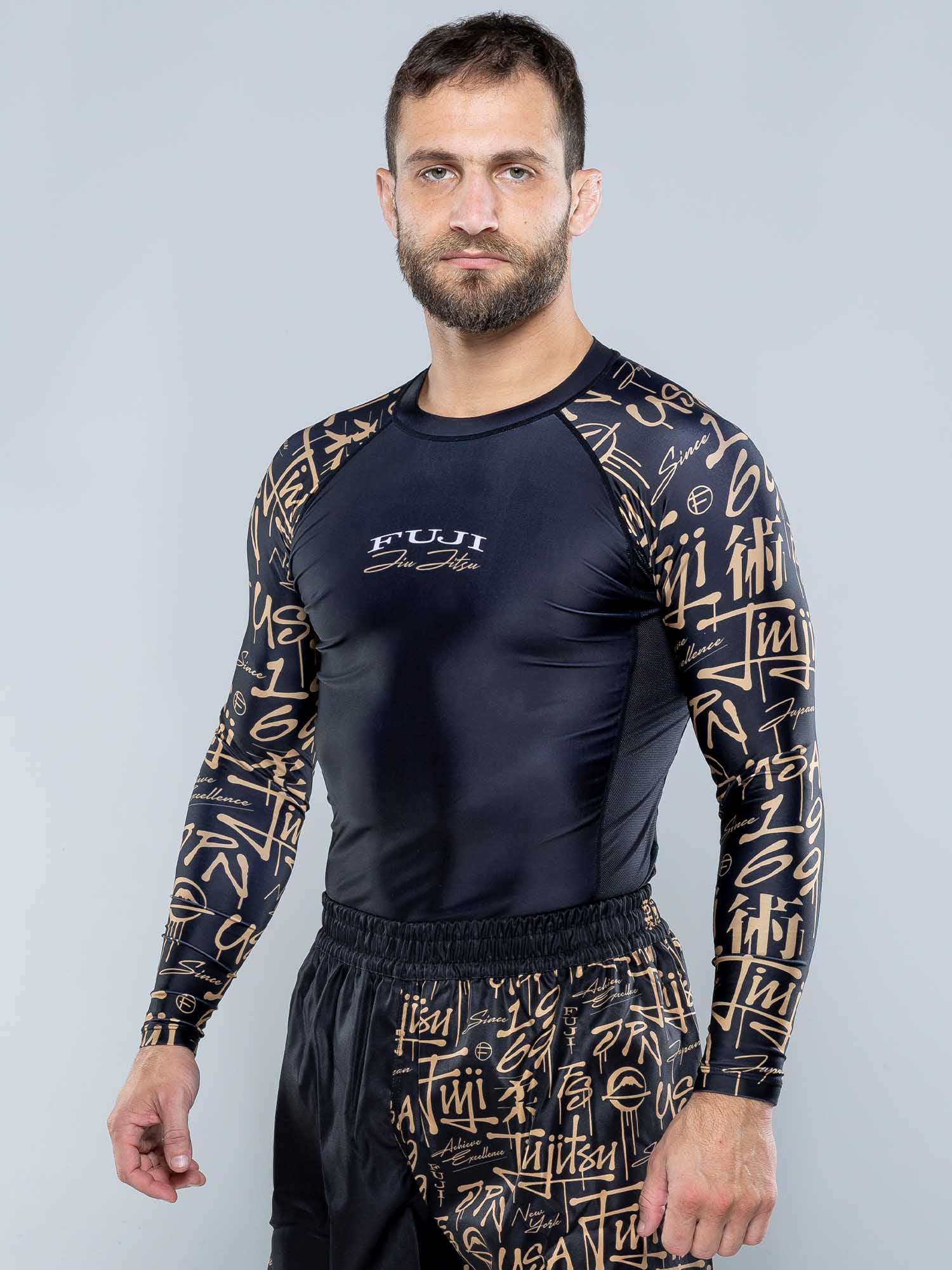 A man with short brown hair and a beard models the Shinobi Long Sleeve Rashguard Black and matching shorts featuring gold graffiti-style writing, standing against a plain gray background.