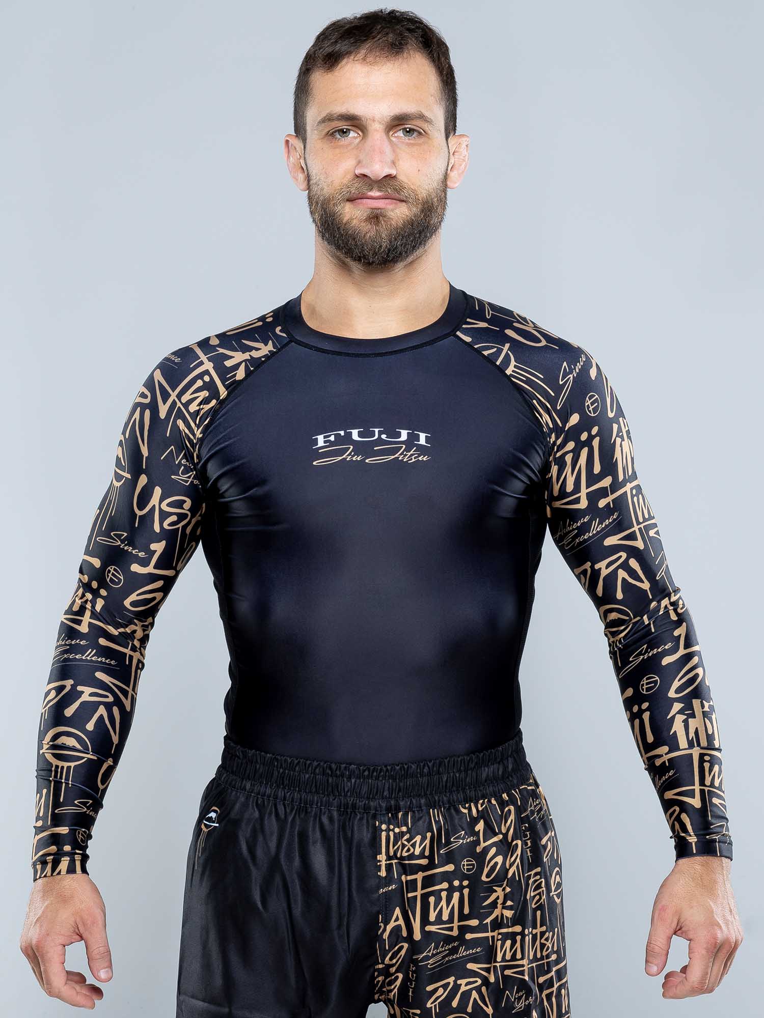A man with short brown hair and a beard wears a black Shinobi Long Sleeve Rashguard and black shorts, standing against a plain gray background.