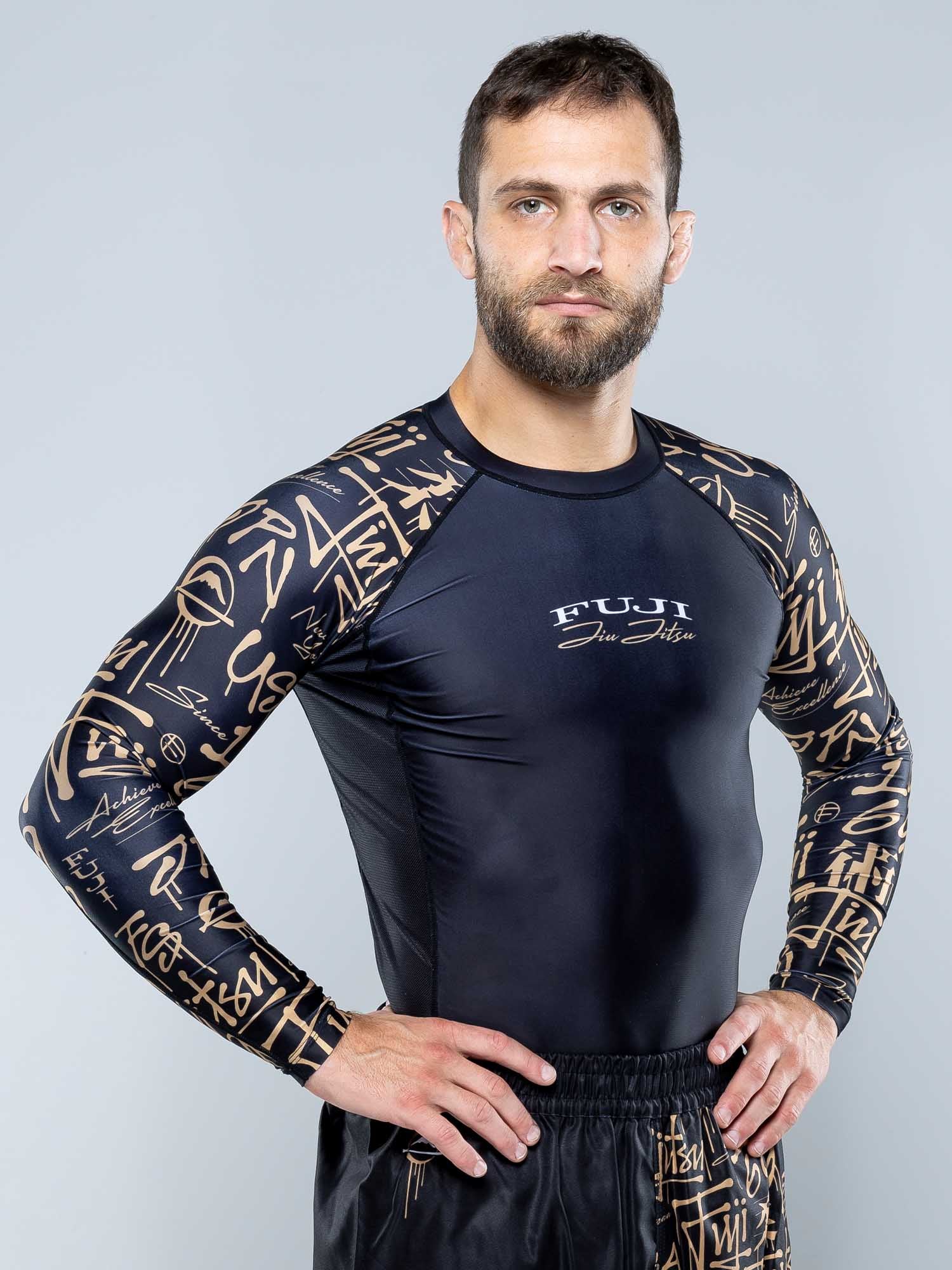 A man with short brown hair and a trimmed beard stands confidently, hands on hips, wearing the Shinobi Long Sleeve Rashguard Black featuring gold sleeve graphics, set against a plain light gray background.