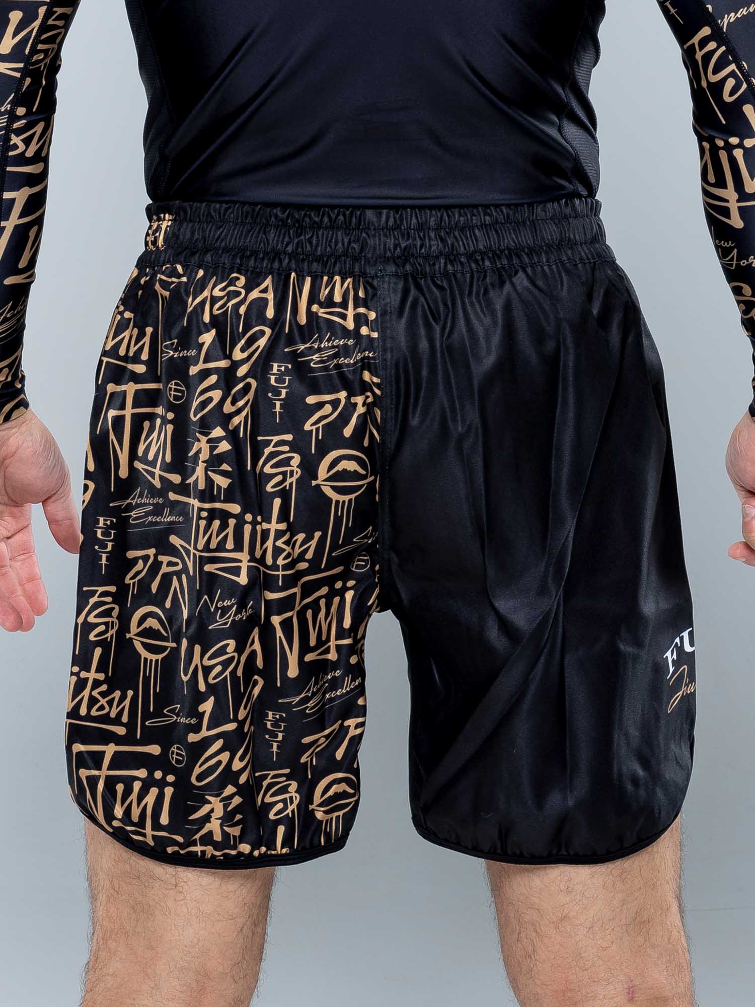 A person wears Shinobi Fight Shorts Black, featuring tan graffiti-style writing and symbols on one side, paired with a matching long-sleeve top. The image highlights the shorts and the person's lower torso and legs.