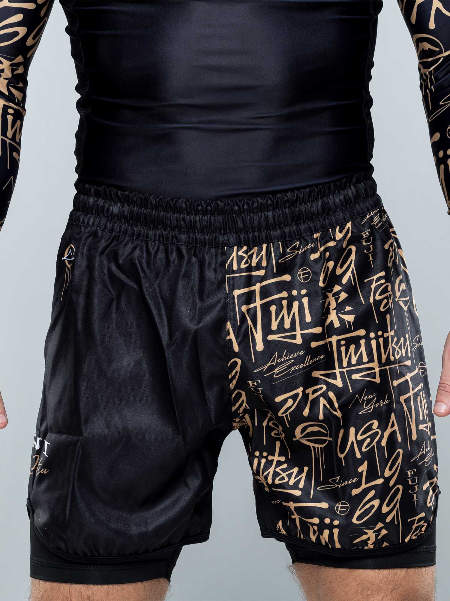 A person wearing Shinobi Fight Shorts Black With Liner, featuring gold graffiti-style writing, shown from the waist to mid-thigh.