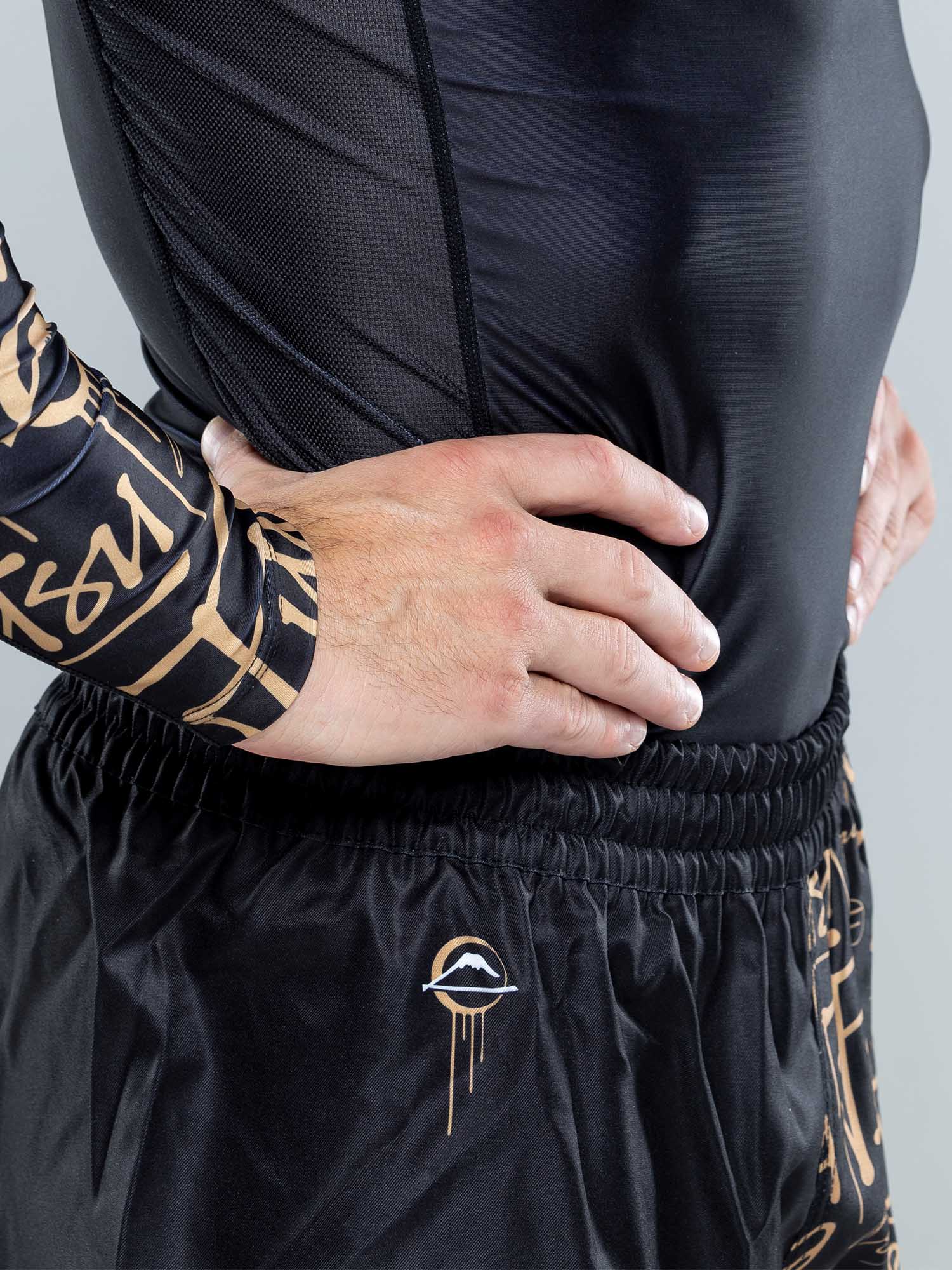 A person wears black athletic clothing with gold patterns. The top has mesh panels, and the Shinobi Fight Shorts Black feature a small stylized eye logo with a line underneath.