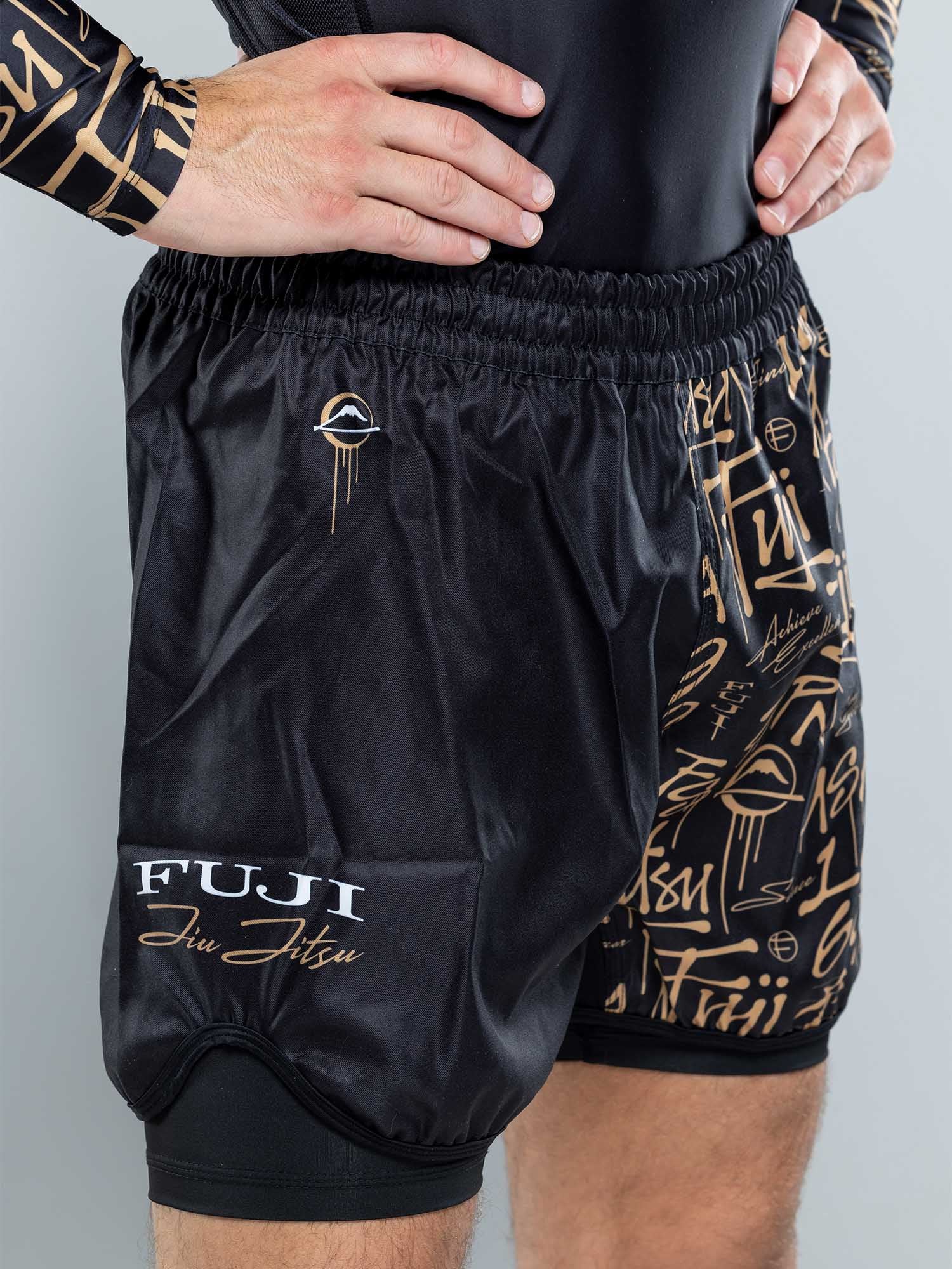 A person wears Shinobi Fight Shorts Black With Liner, featuring gold patterns and logos, standing with one hand on their hip against a plain, light-colored background.