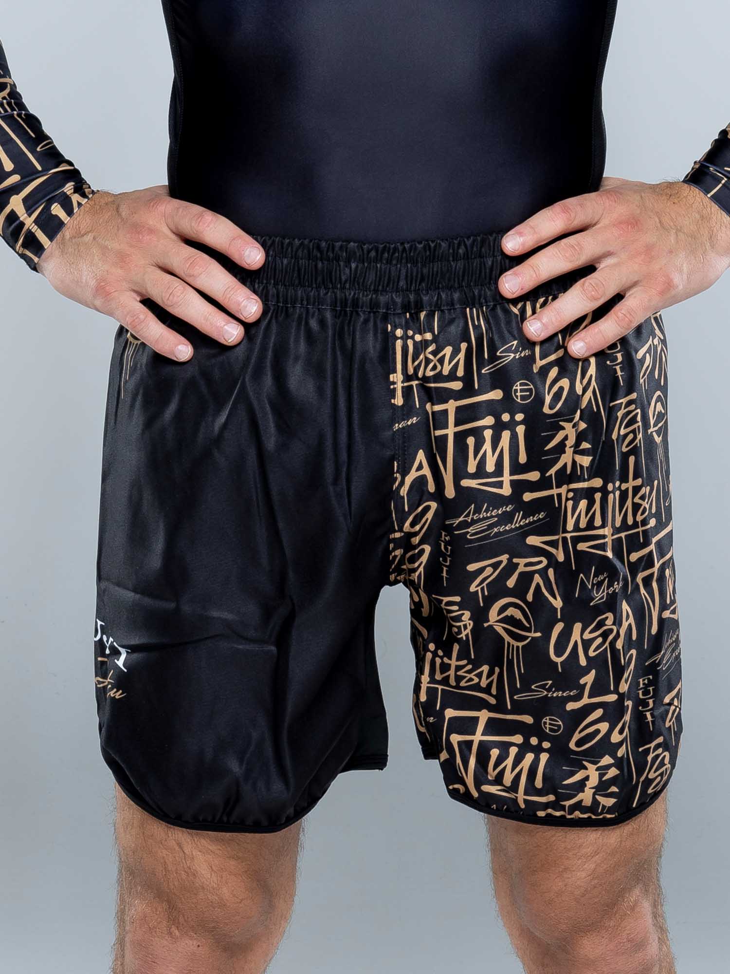A person models Shinobi Fight Shorts Black, featuring gold graffiti-style writing, with hands on hips. Only the torso and upper legs are visible against a plain, light-colored background.