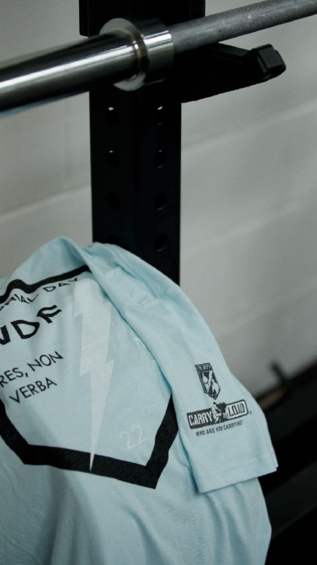 A light blue t-shirt with printed graphics, including a lightning bolt and text, draped over a barbell rack in a gym setting. The shirt also features a Carry The Load patch on the sleeve.
