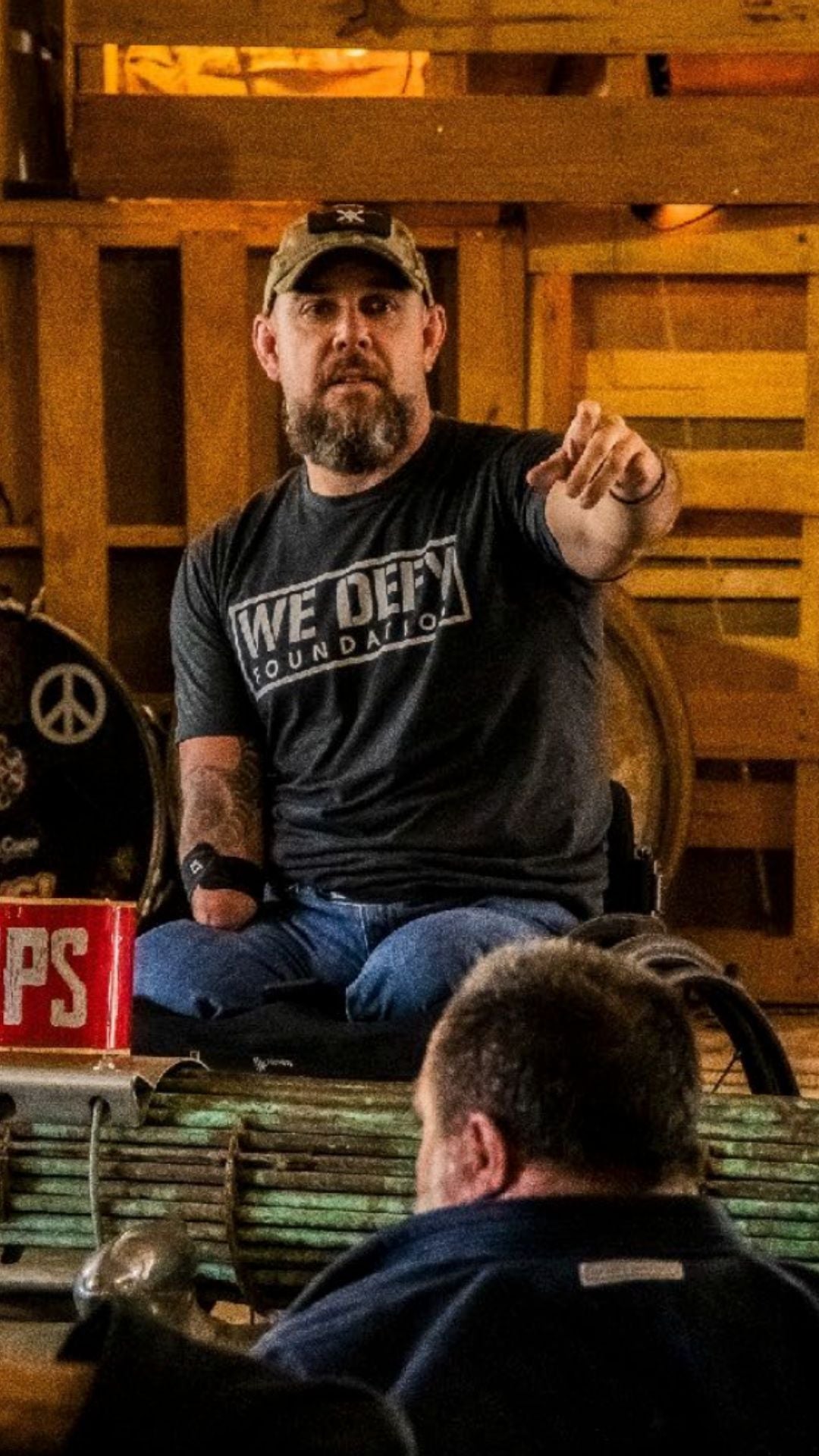 A bearded man wearing a We Defy Foundation t-shirt and a baseball cap gestures with his right hand while seated indoors. He is an amputee, missing his left arm below the elbow. Another person is visible in the foreground.