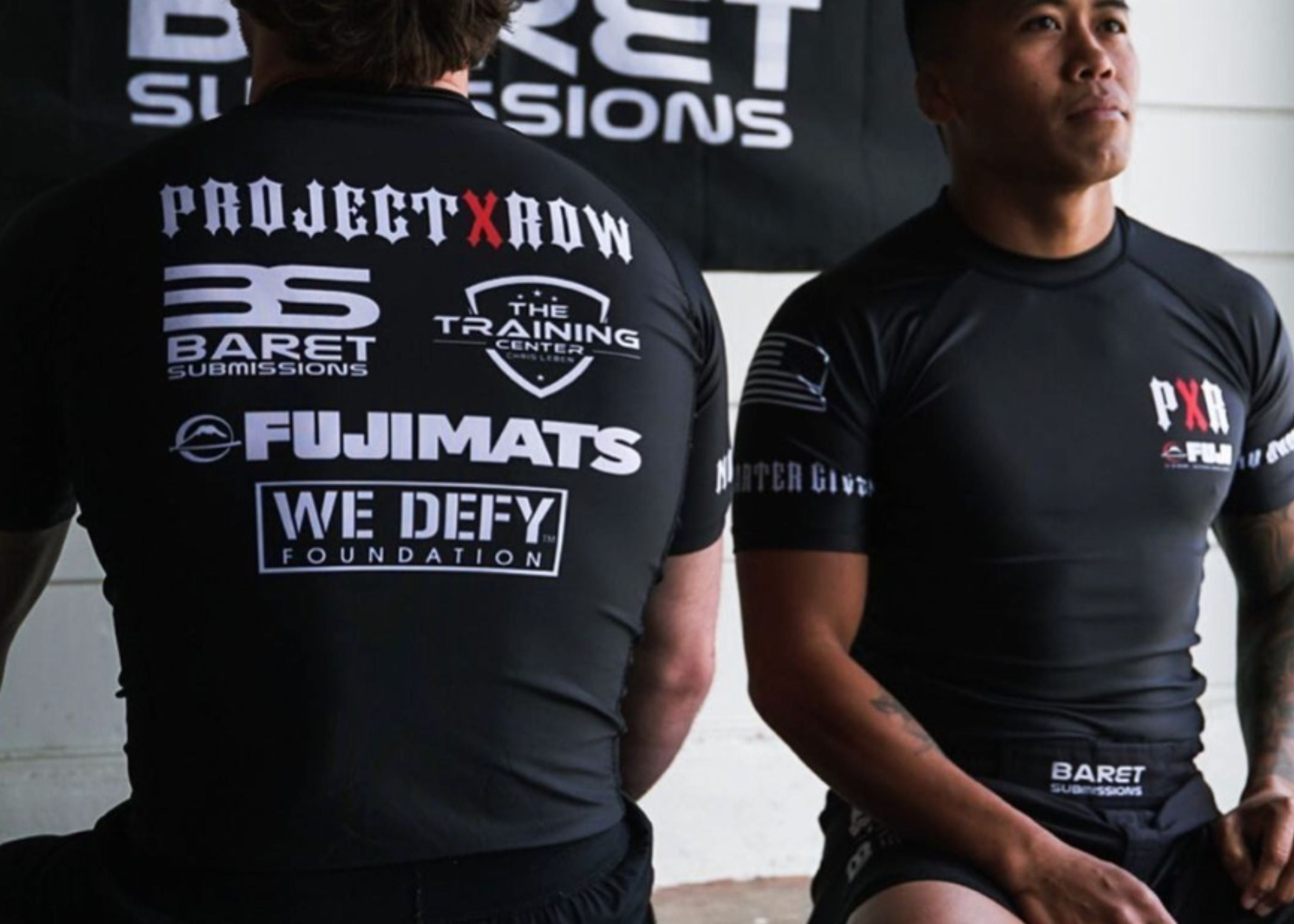 Two men in black athletic shirts sit side by side. The back of one shirt displays various logos, including Project X Row, Baret Submissions, and Fujimats. The other man faces forward.