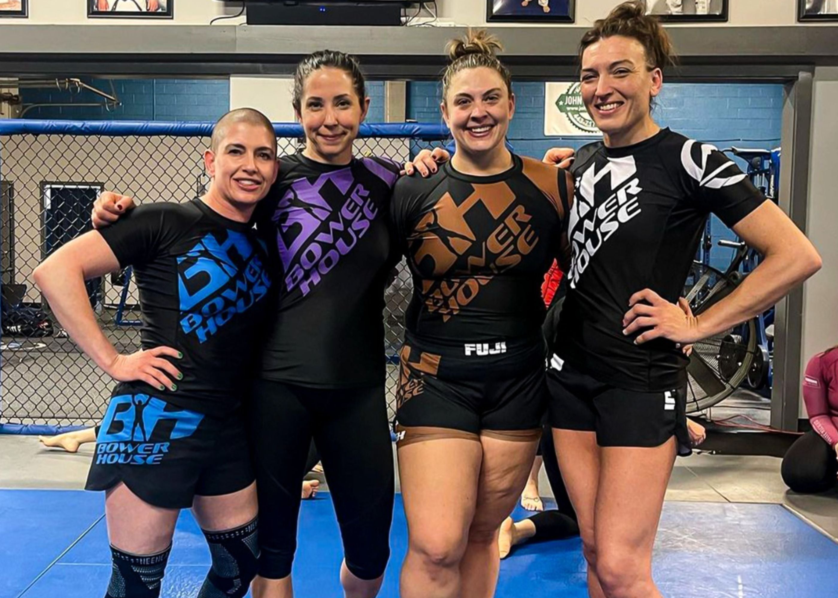 Four women in grappling attire stand together smiling inside a gym, with blue gym mats and a fenced cage wall behind them. They each wear matching Power House rash guards and shorts in different colors.