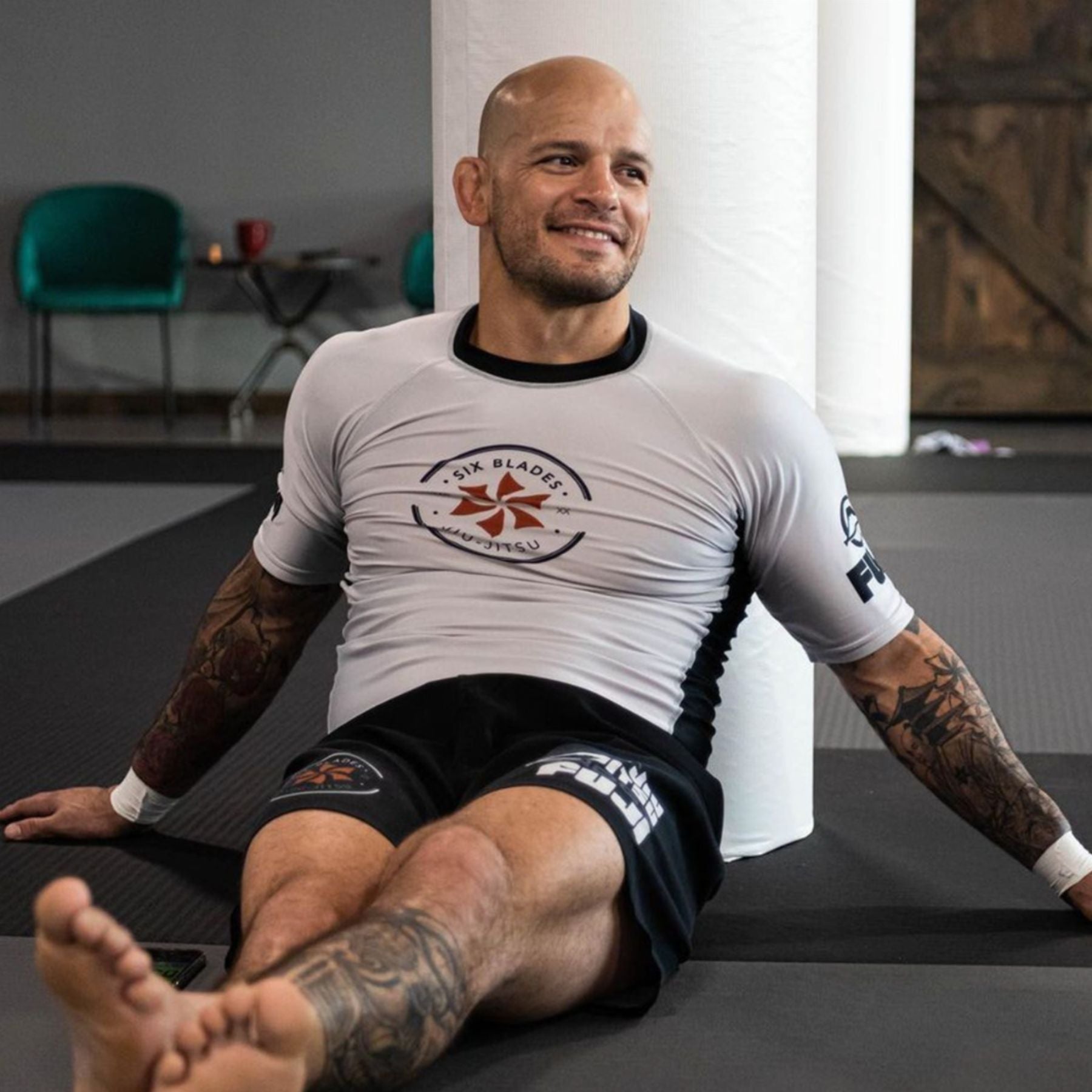 A smiling, bald man in a white rash guard and black shorts with tattoos on his arms sits on a gym mat, leaning against a padded pole in a martial arts training facility.