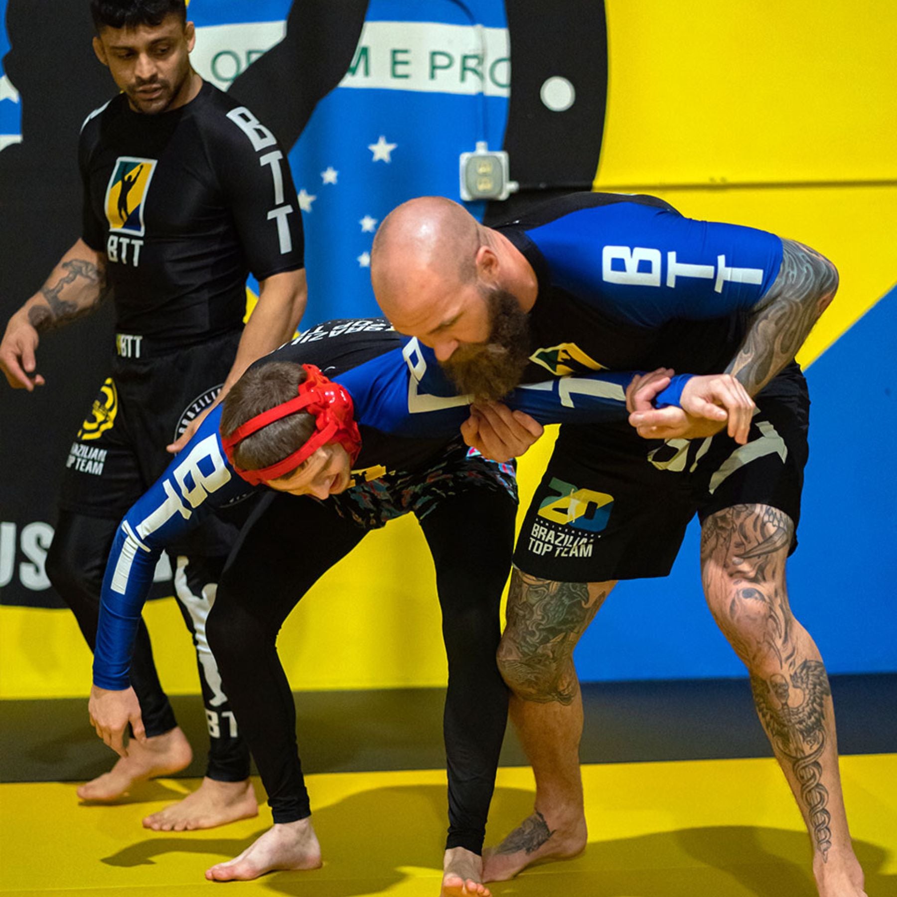 Three people practice Brazilian Jiu-Jitsu in a gym. One person demonstrates a grappling move on another, while a third person observes. They all wear matching rash guards and shorts. The background features a bright yellow wall.