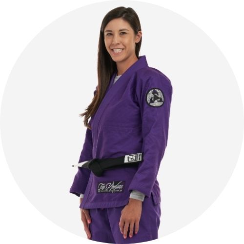A woman with long brown hair smiles while wearing a purple Brazilian jiu-jitsu gi with a black belt, standing against a plain light background.