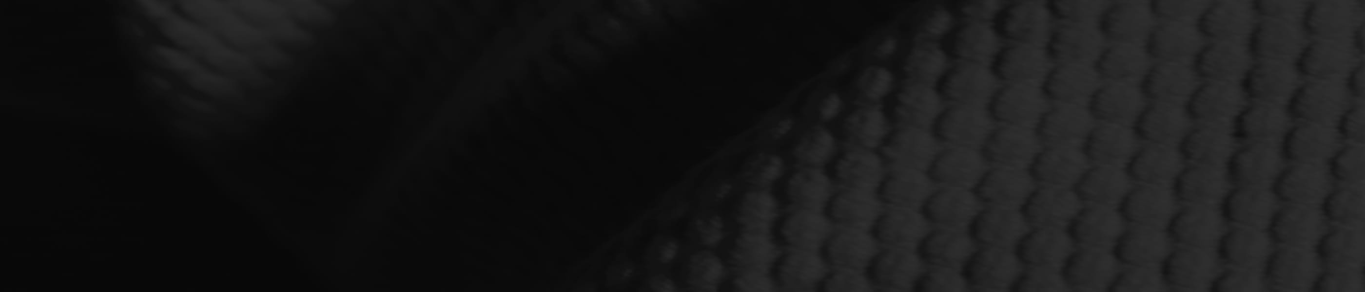 Close-up of a dark, textured fabric with a repeating, slightly raised diamond or hexagonal pattern, shown in low lighting with visible shadows creating depth and contrast.