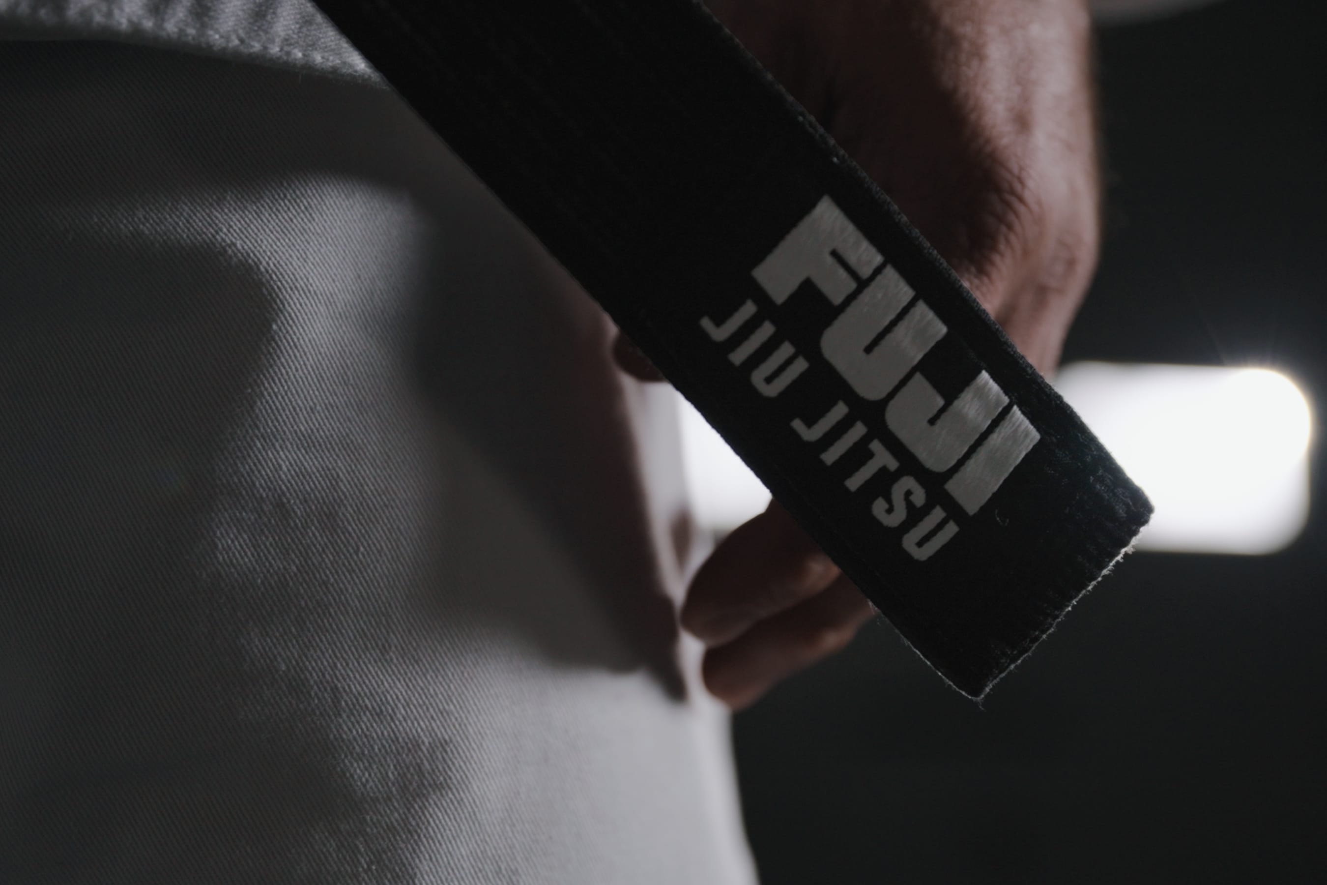 Close-up of a person holding a black belt with the words FUJI JIU JITSU embroidered on it, wearing a white uniform, with a blurred background.