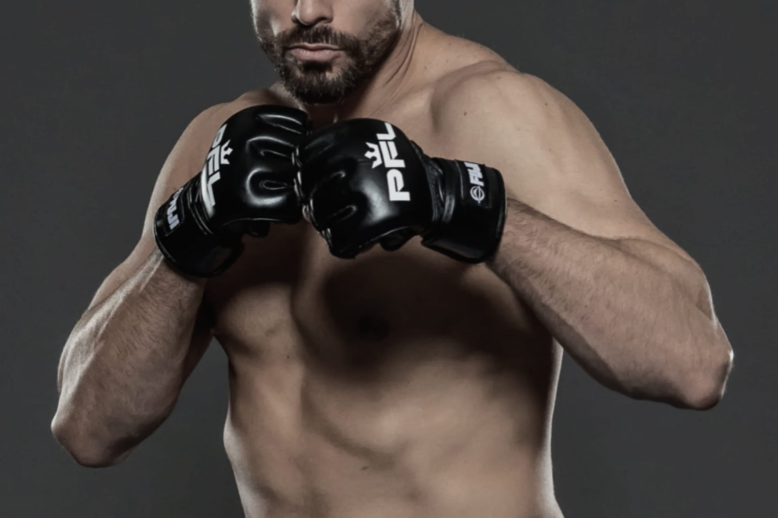 A shirtless male MMA fighter poses with fists raised in front of his chest, wearing black gloves against a dark background.