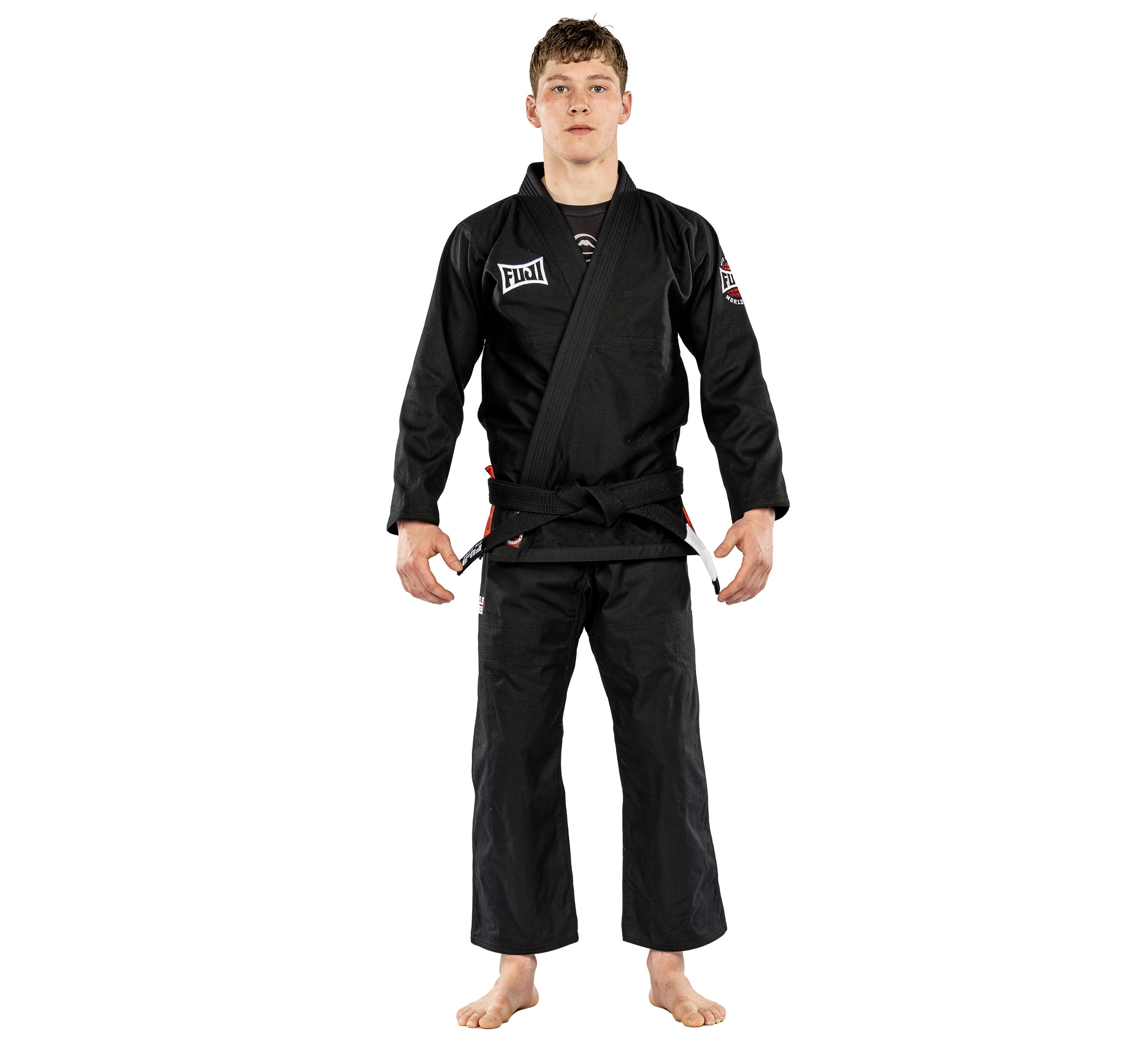 A young man stands barefoot against a plain white background, wearing the Suparaito BJJ Gi Worldwide Edition Black with chest and sleeve patches and a black belt tied at the waist.