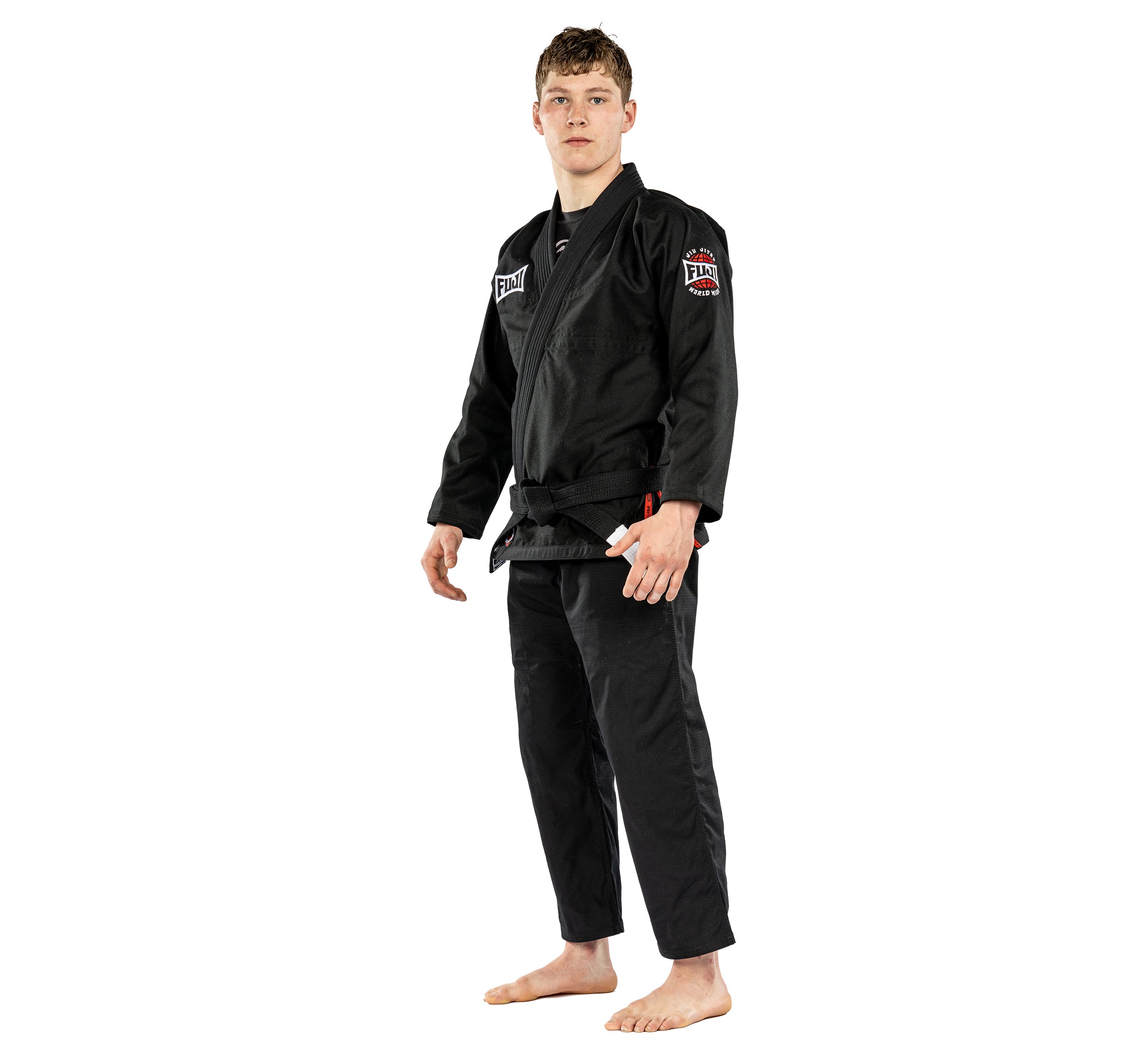 A young man stands barefoot against a plain white background, wearing the Suparaito BJJ Gi Worldwide Edition Black with patches and a black belt.