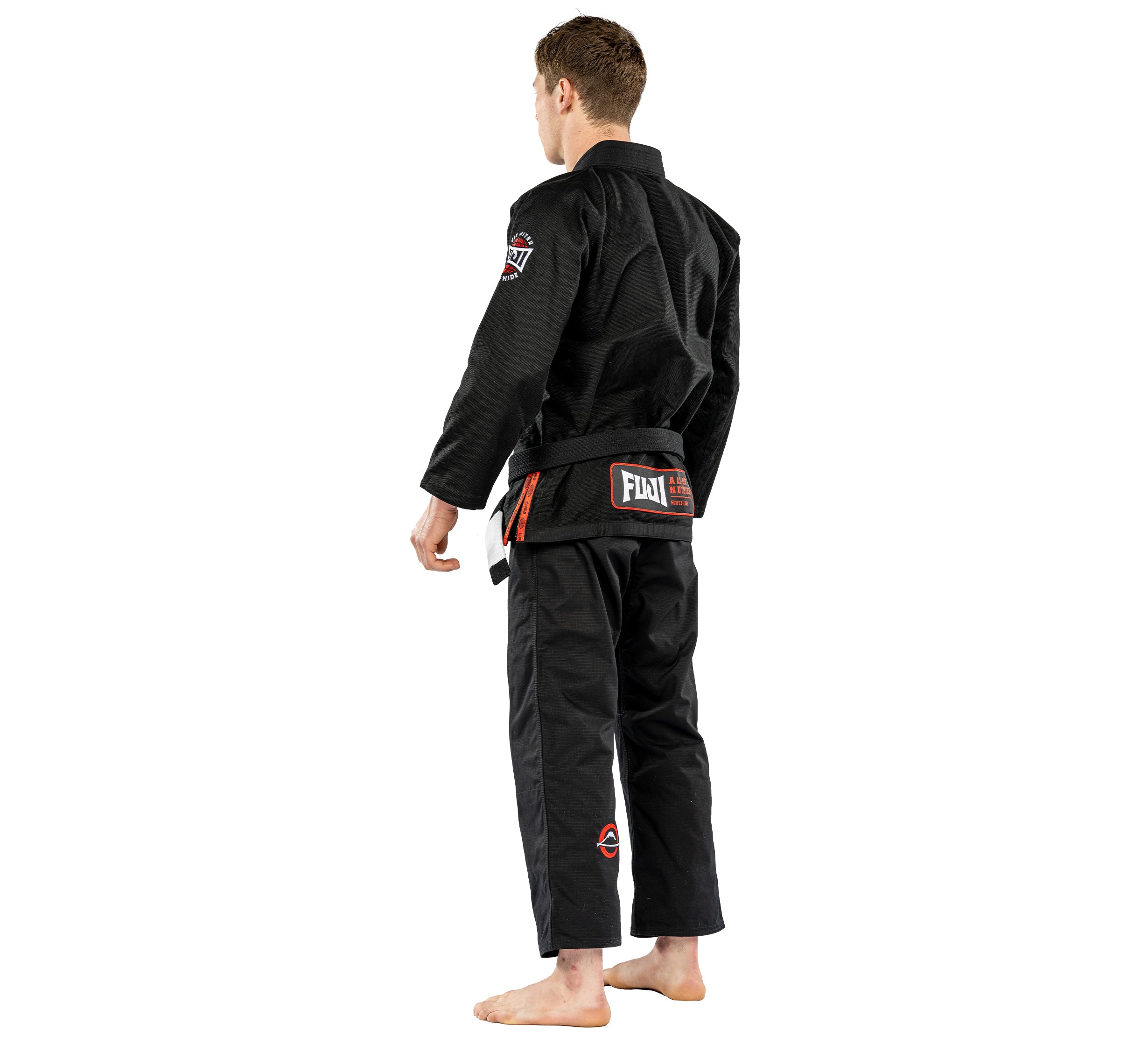 A person wearing the Suparaito BJJ Gi Worldwide Edition Black and a black belt stands barefoot, facing away from the camera against a plain white background.