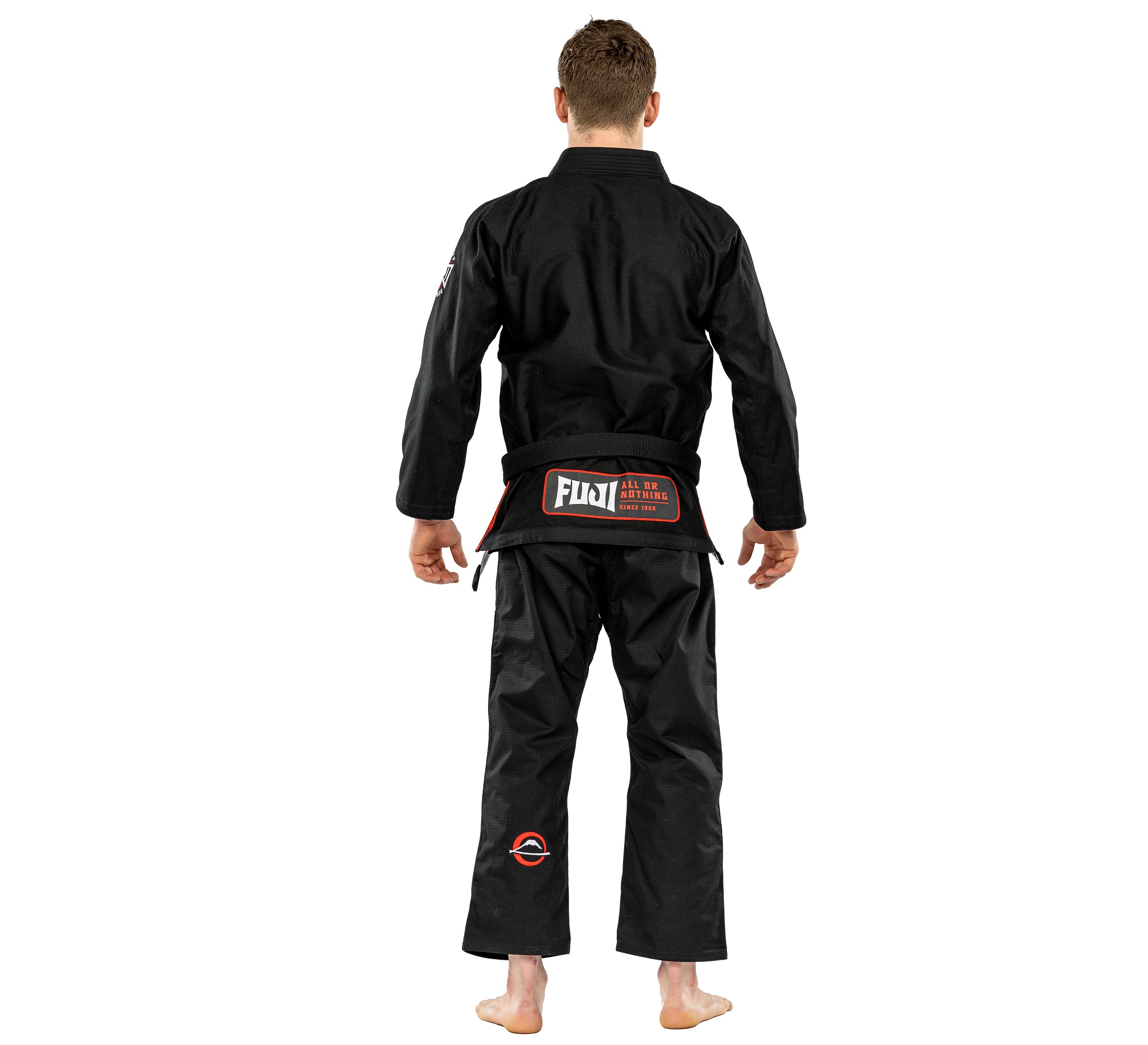 A person stands barefoot facing away, wearing the Suparaito BJJ Gi Worldwide Edition Black with "FUJI" on the back of the belt and small red and white patches on the uniform.