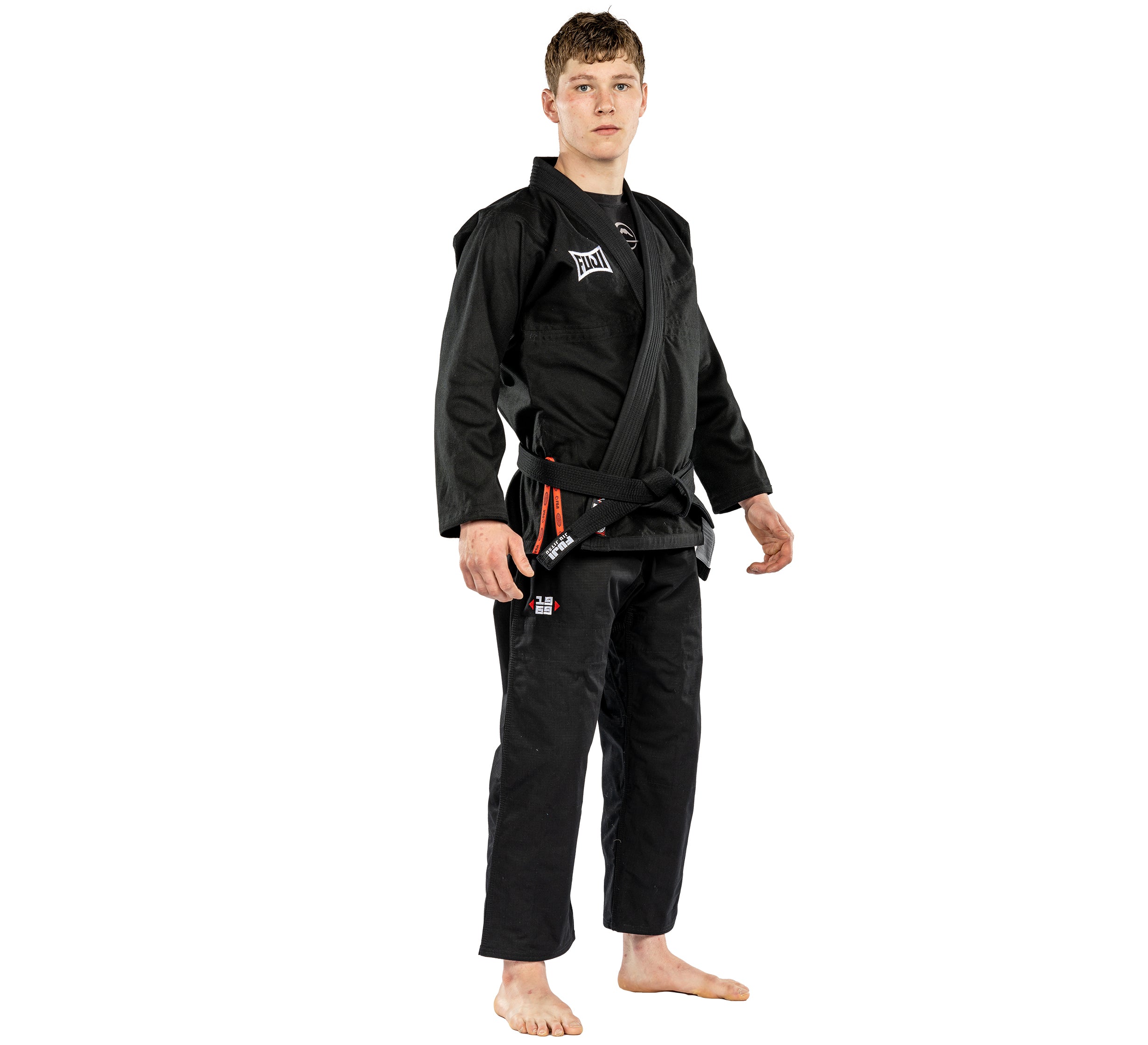 A young man stands barefoot in the Suparaito BJJ Gi Worldwide Edition Black with a black belt, looking forward with a neutral expression against a plain white background.