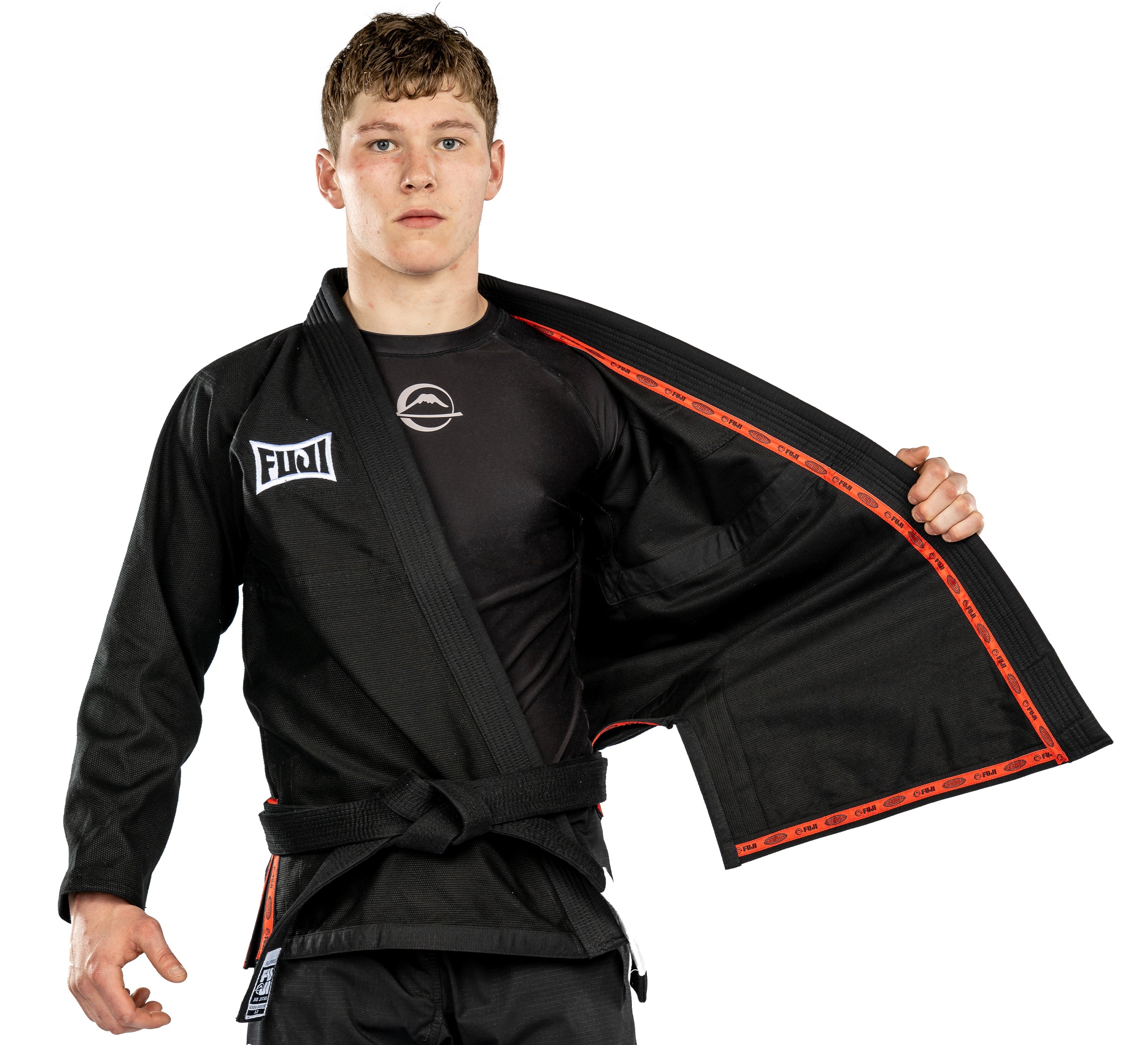 A young man in the Suparaito BJJ Gi Worldwide Edition Black with patches and a black belt stands facing forward, holding open one side of his gi to reveal a rash guard underneath.