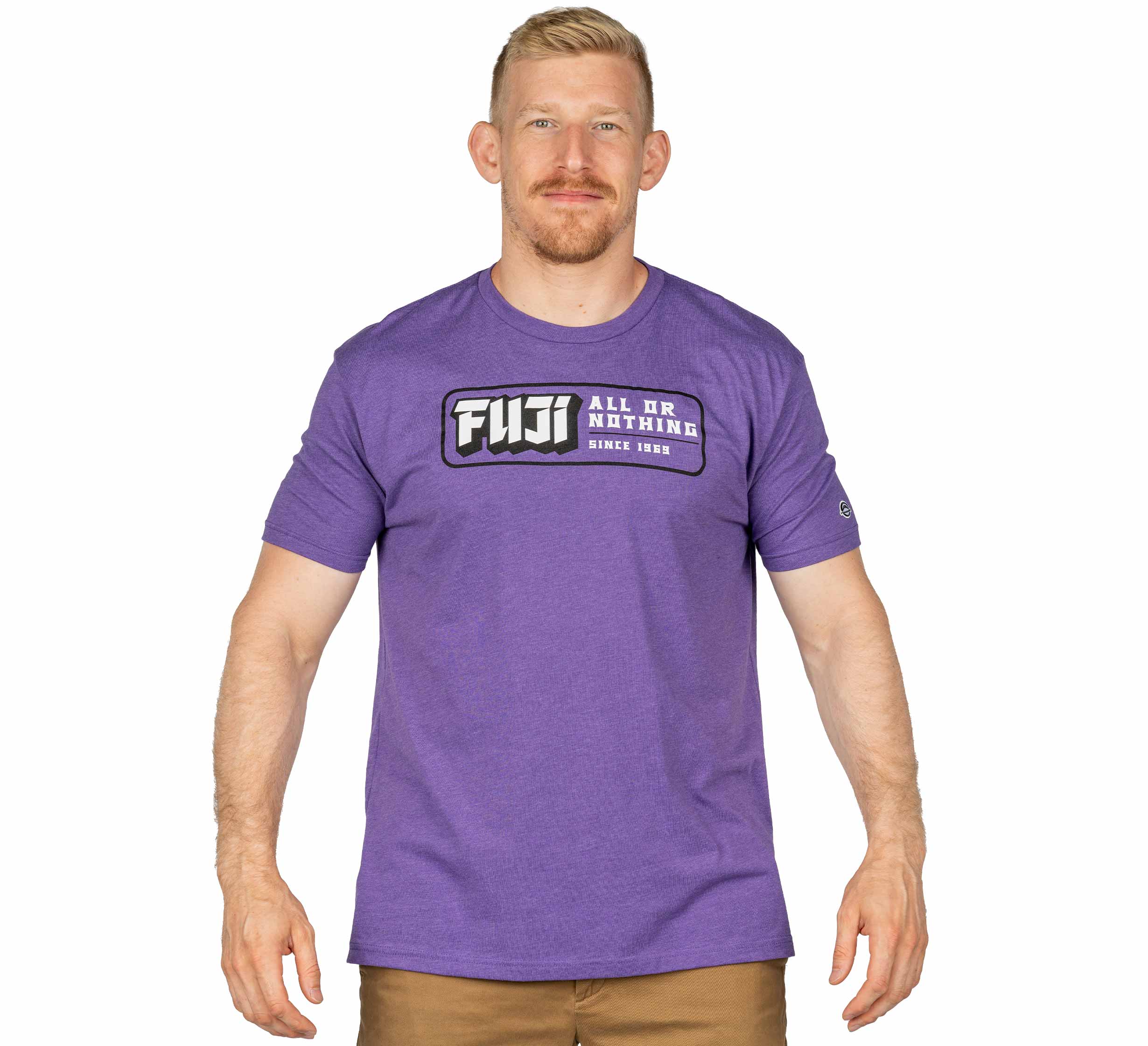 A man with short blond hair and a beard is standing, facing forward and smiling slightly. He is wearing khaki pants and a purple Ranked Jiu-Jitsu T-Shirt that says FUJI ALL OR NOTHING SINCE 1969.
