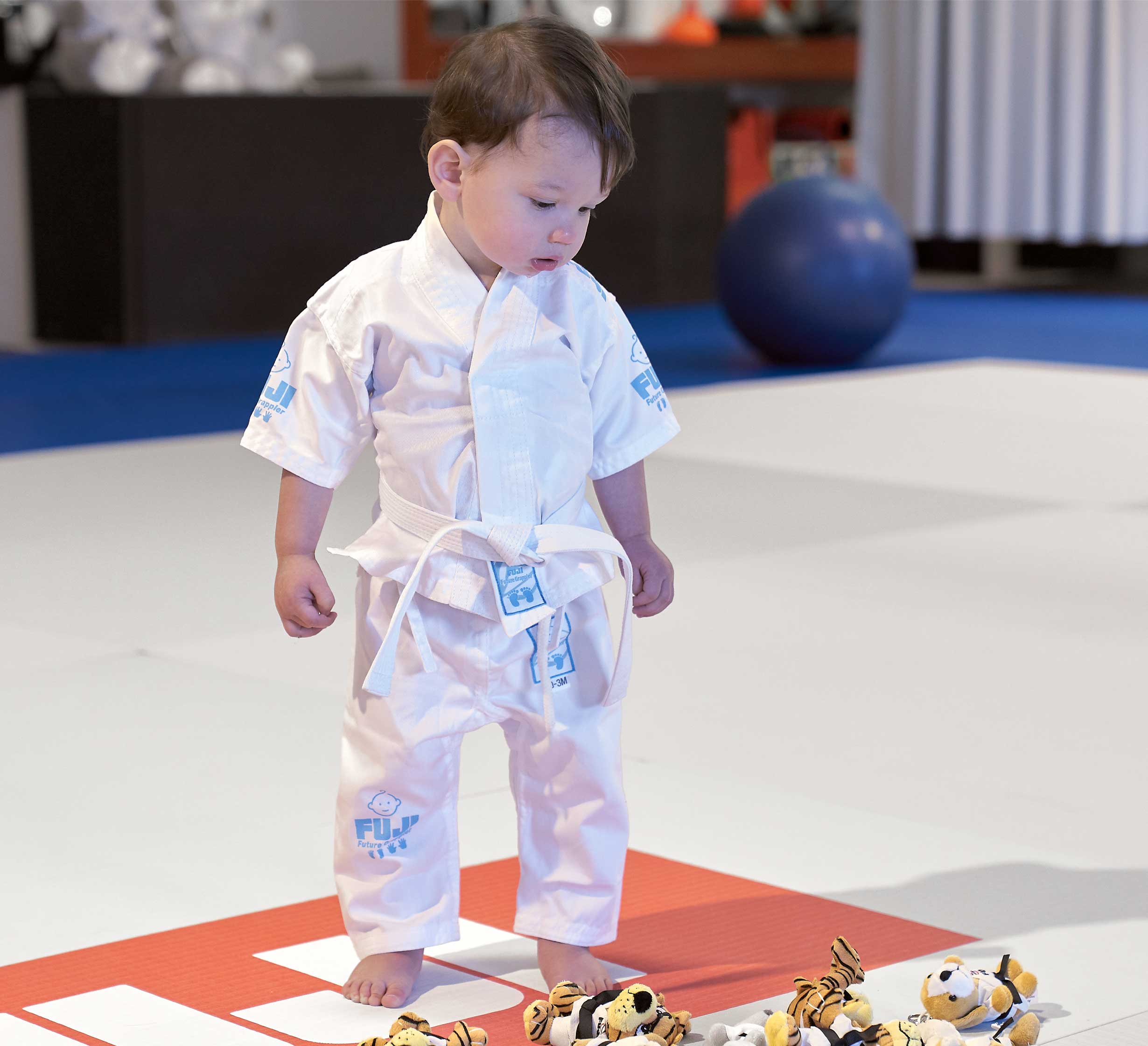 A young child wearing the Baby Gi Blue martial arts uniform stands barefoot on a mat, looking down at a row of plush toys. A blue exercise ball and gym equipment can be seen in the background.