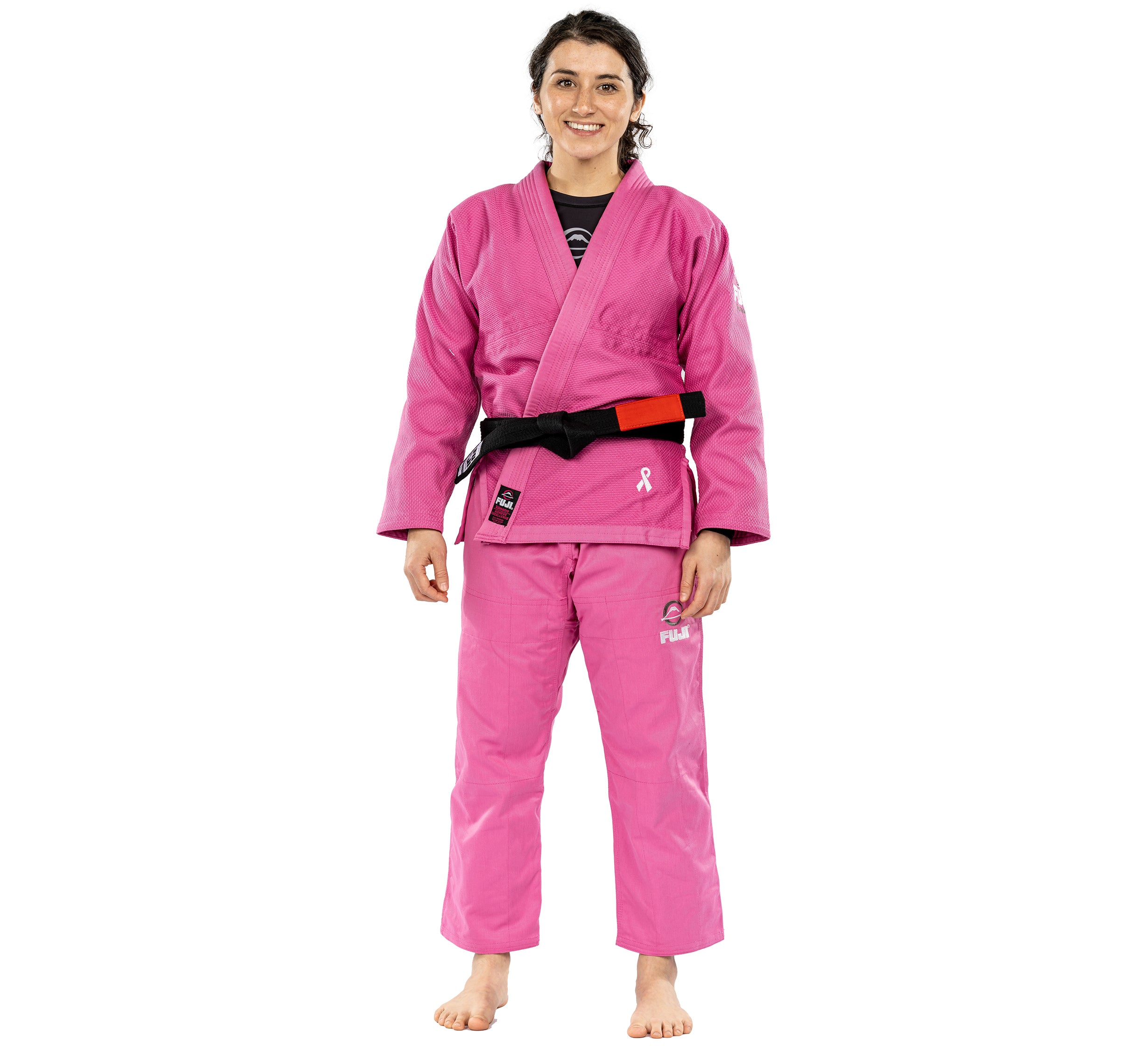 A smiling person stands barefoot in the All Around Womens BJJ Gi Pink, featuring white ribbon breast cancer awareness logos on the chest and leg and a black-red belt. The background is plain white.
