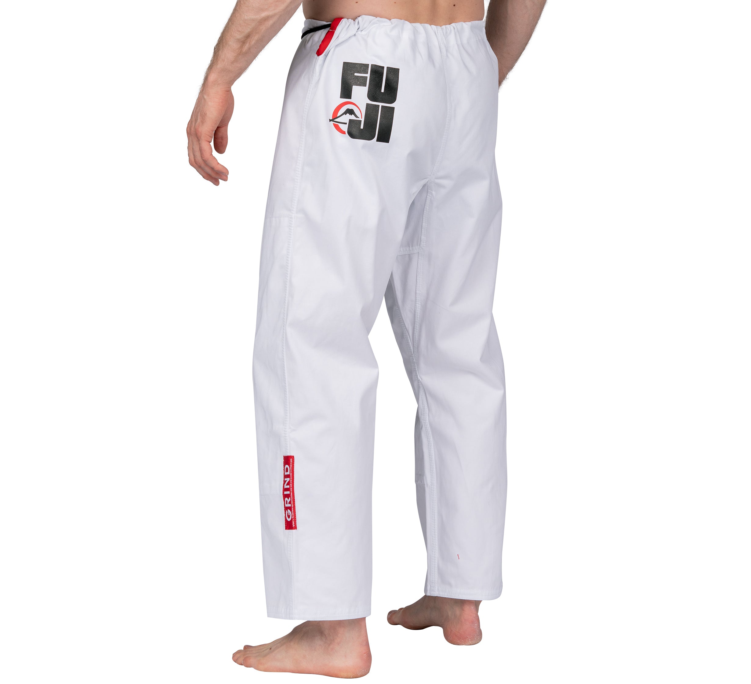 A person stands barefoot, wearing Elemental BJJ Gi Grind White pants with a Fuji logo and red Shinju patch. The image focuses on the back and side of the pants, showing only their torso and legs.