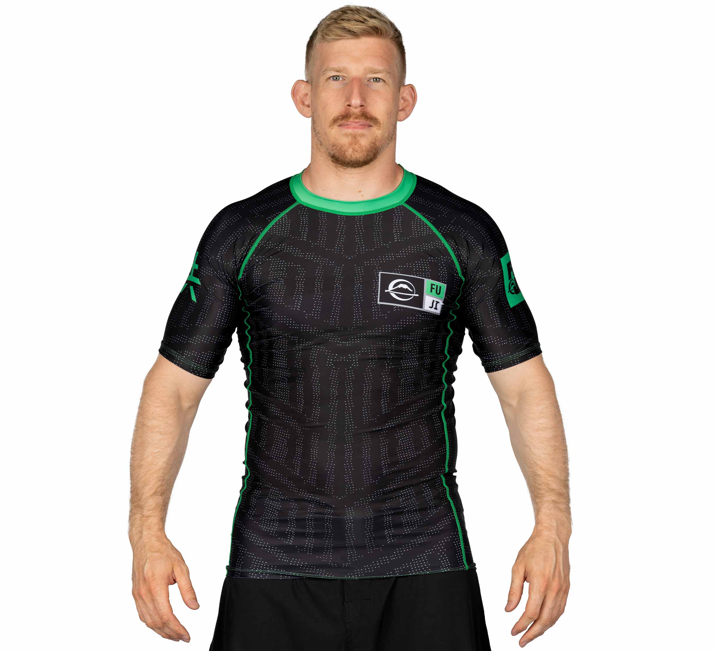 A man with short blond hair and a beard wears the Team FUJI Short Sleeve Rashguard Green, featuring logos on the chest and sleeves, while standing against a white background.