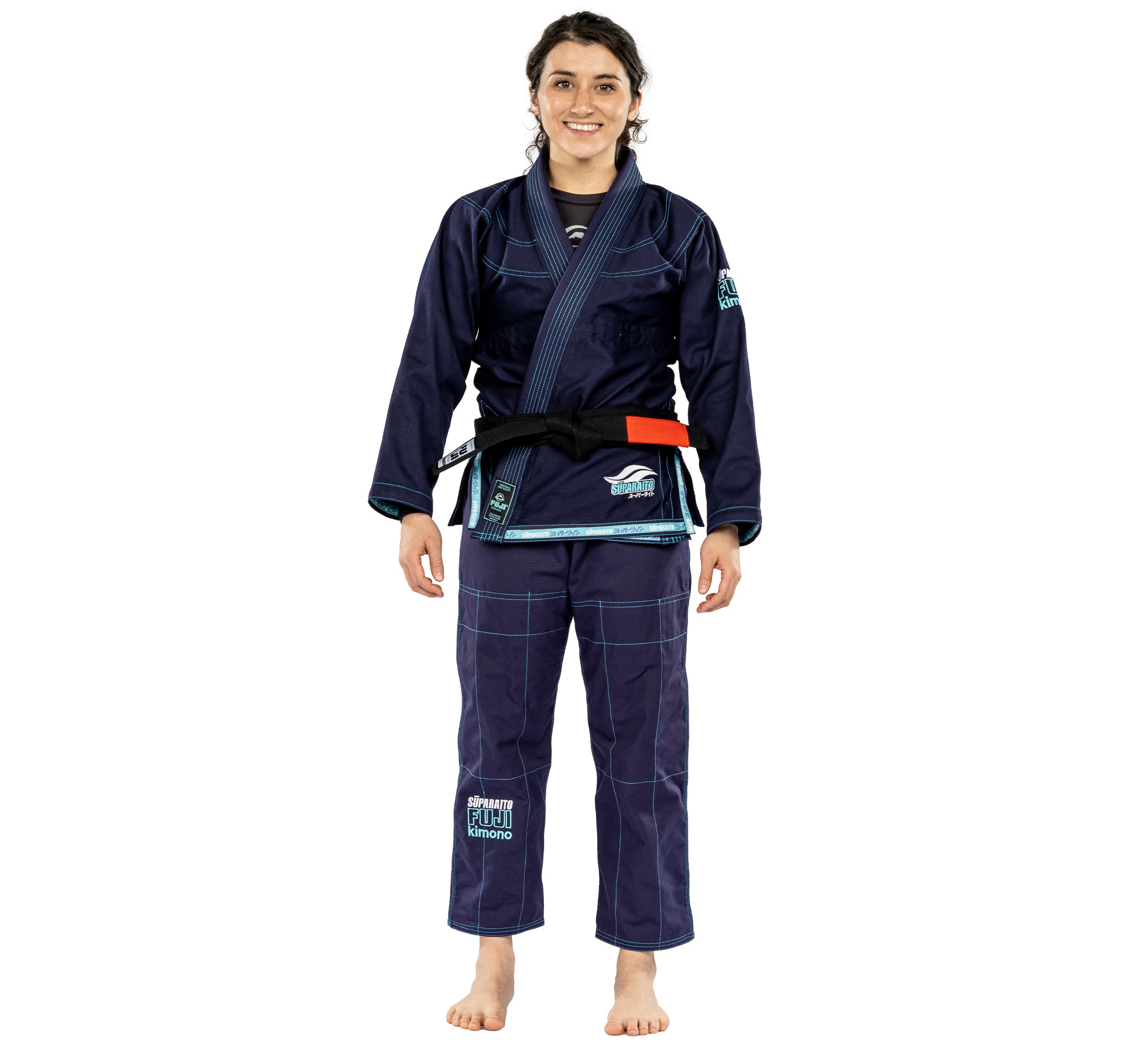 A smiling woman stands barefoot in the Suparaito Womens BJJ Gi Navy, featuring a black belt with red rank bar. She faces forward with her hands relaxed at her sides.