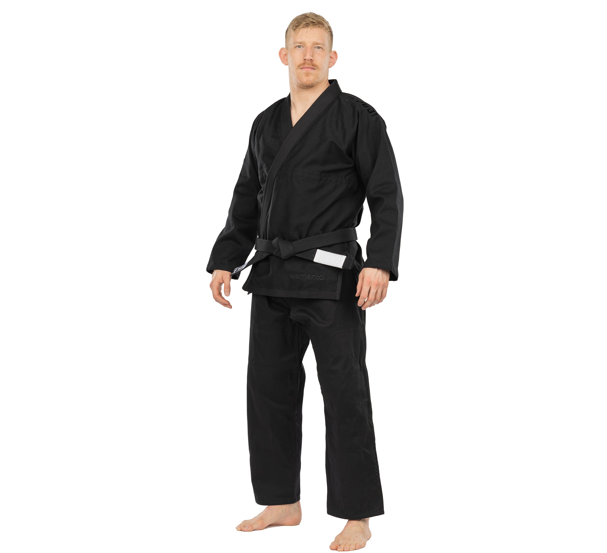 A man stands barefoot in the Elemental BJJ Gi Black with a black belt, looking forward with a neutral expression against a plain white background.