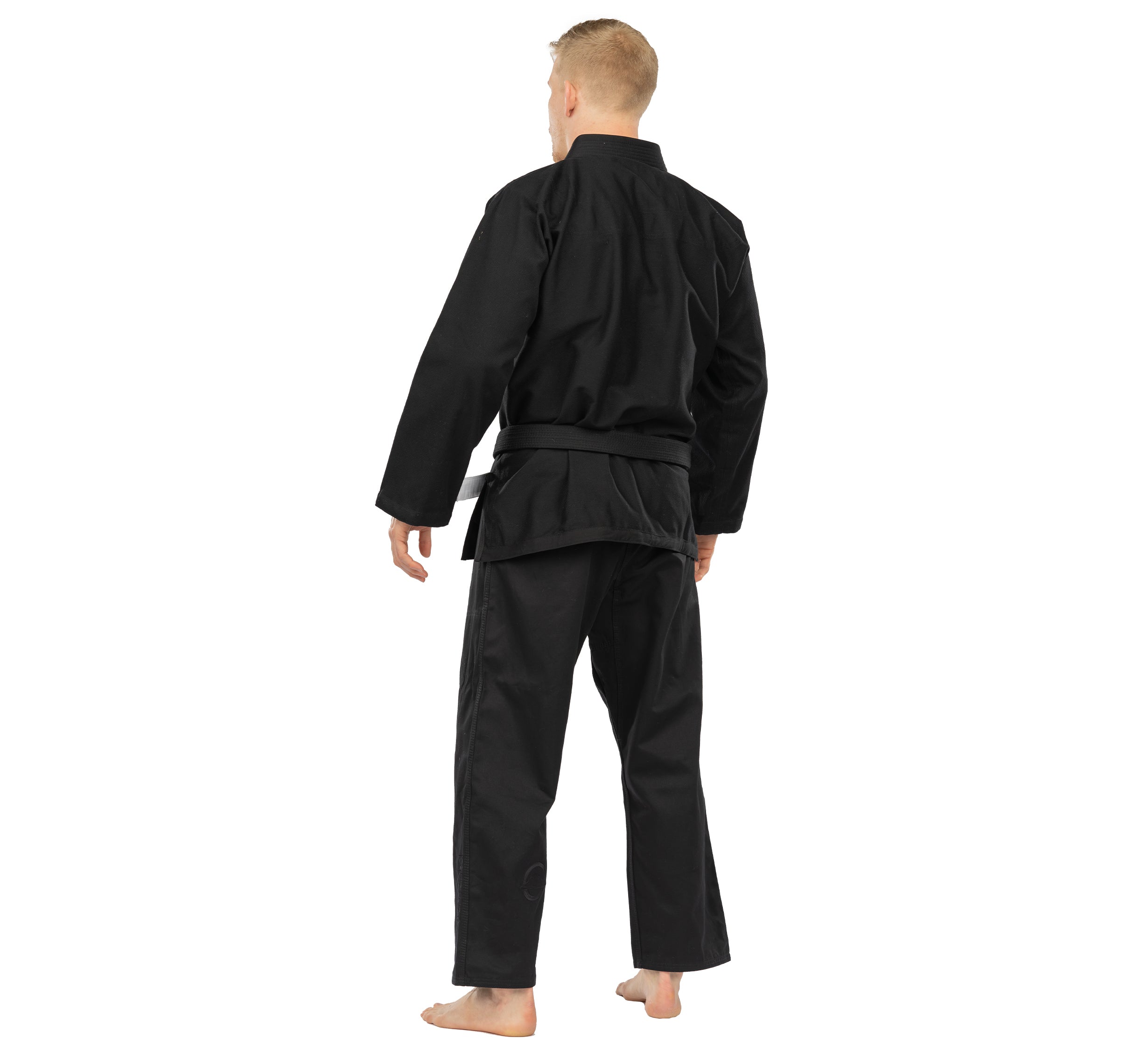 A barefoot individual with short blond hair, wearing the Elemental BJJ Gi Black and a black belt, stands facing away on a white background.