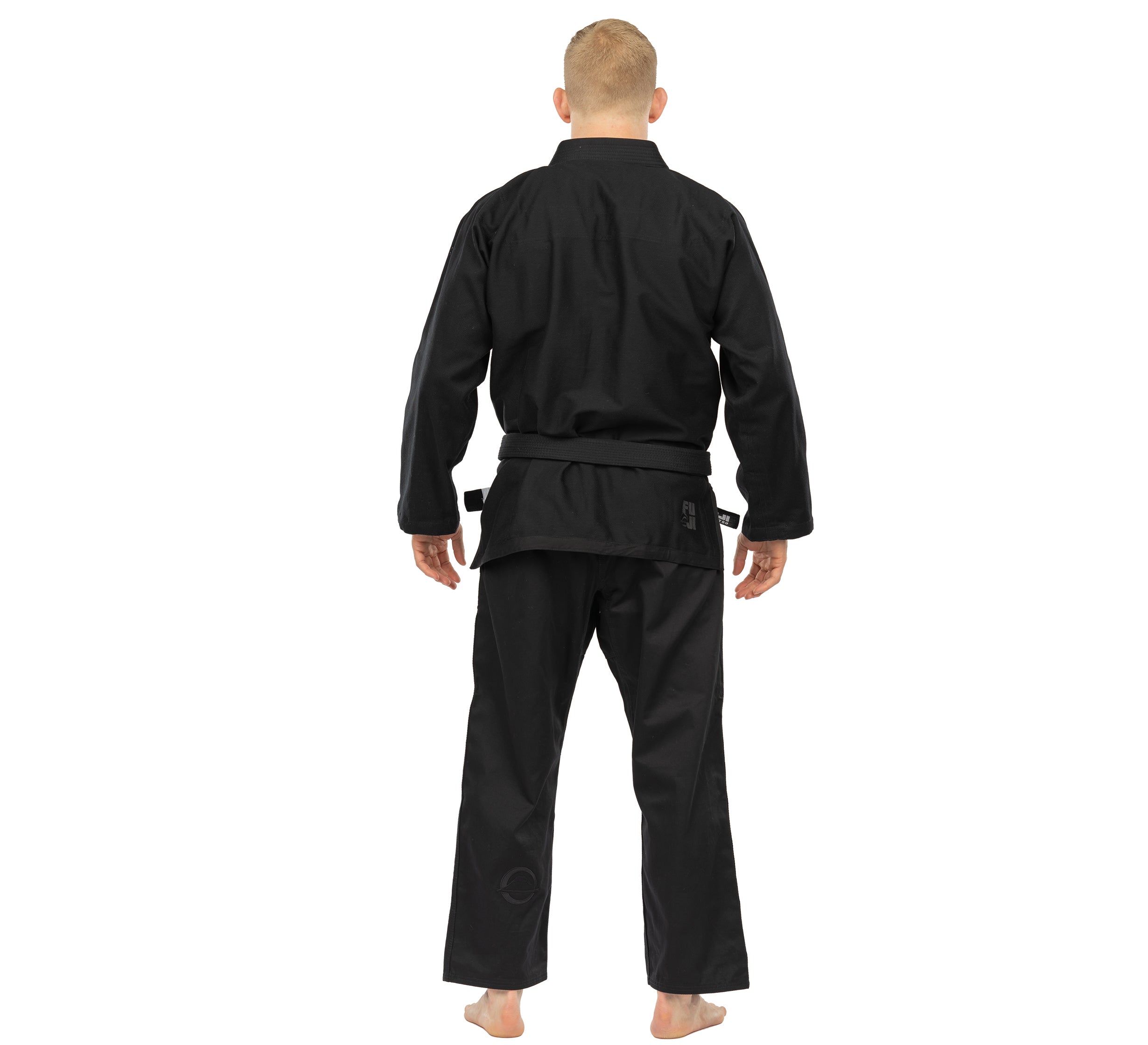 A person with short hair stands barefoot, facing away, wearing the Elemental BJJ Gi Black with a belt against a plain white background.