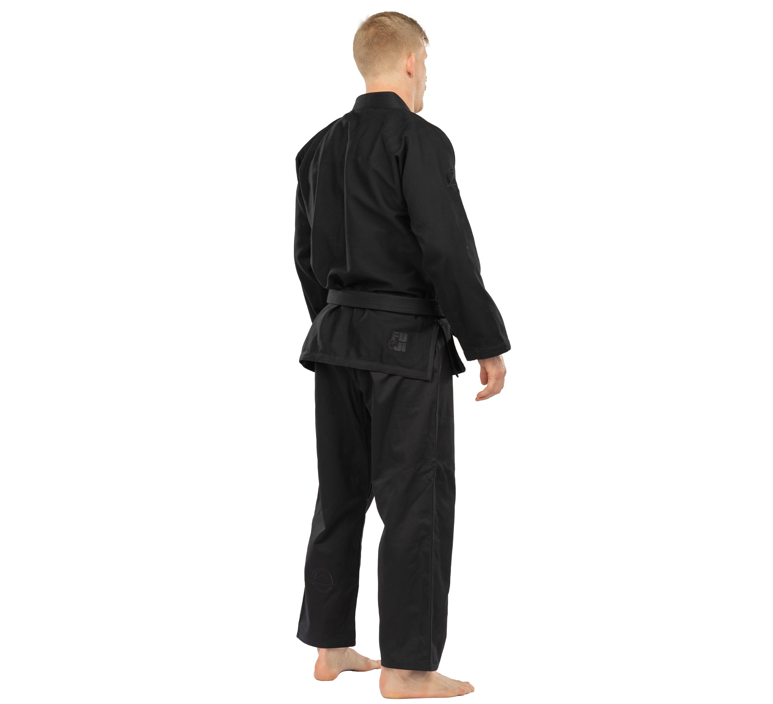 A person with short blond hair stands barefoot, facing away, wearing the Elemental BJJ Gi Black and a black belt against a plain white background.