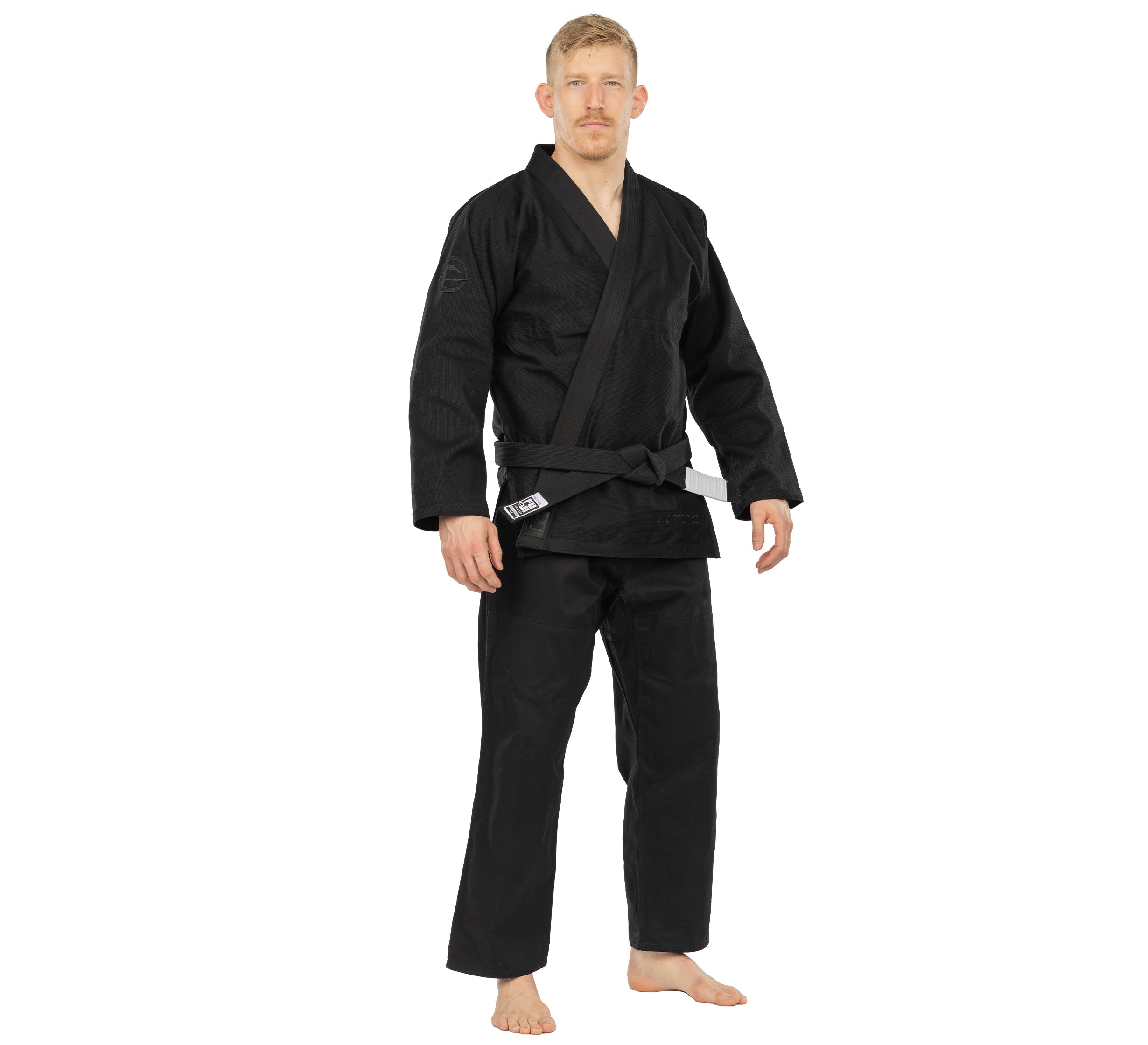 A man stands barefoot facing forward against a plain white background, wearing the Elemental BJJ Gi Black with a black belt.