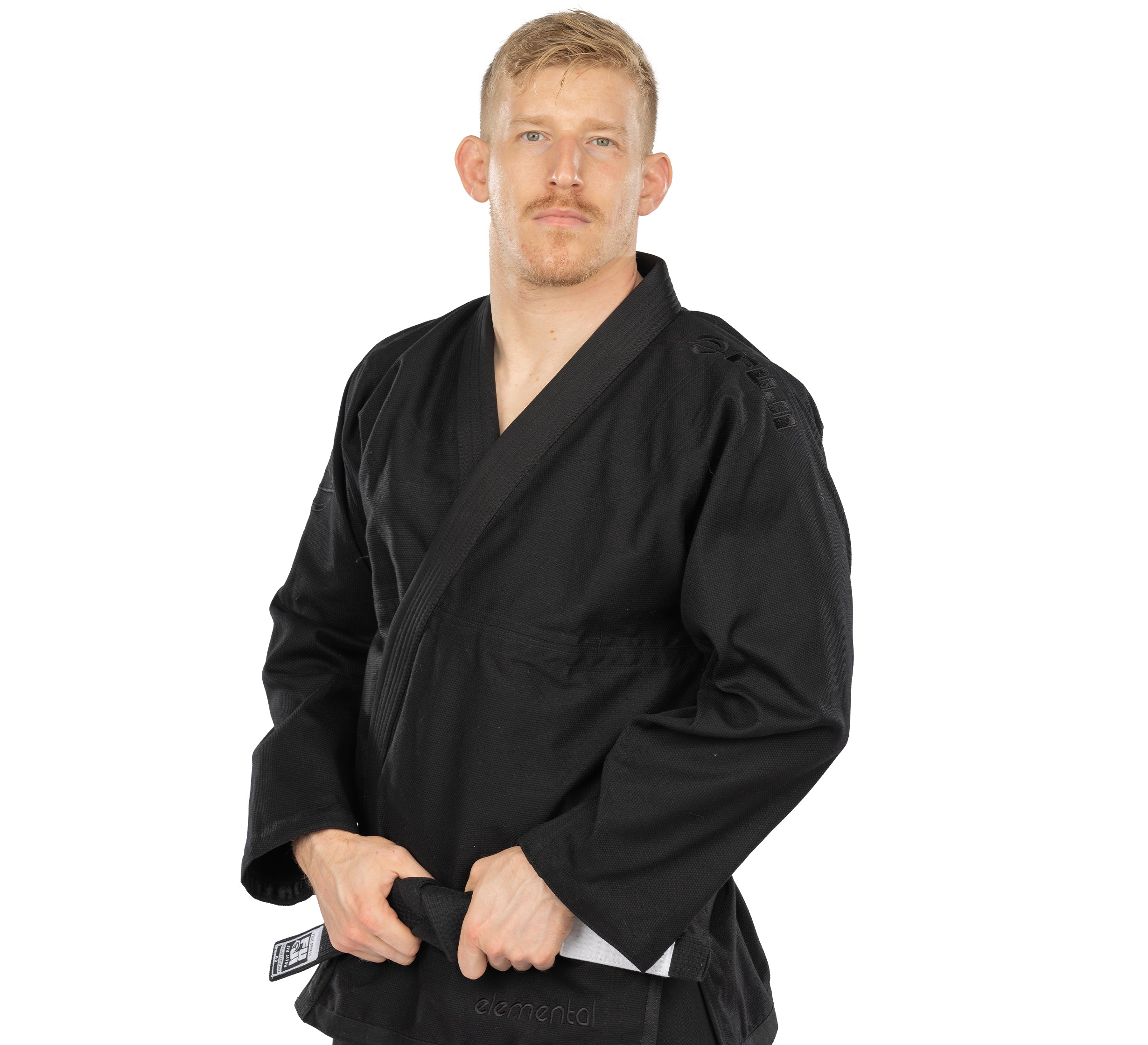A man with short blond hair and a mustache wears an Elemental BJJ Gi Black, holding the ends of his black belt while standing against a plain white background.