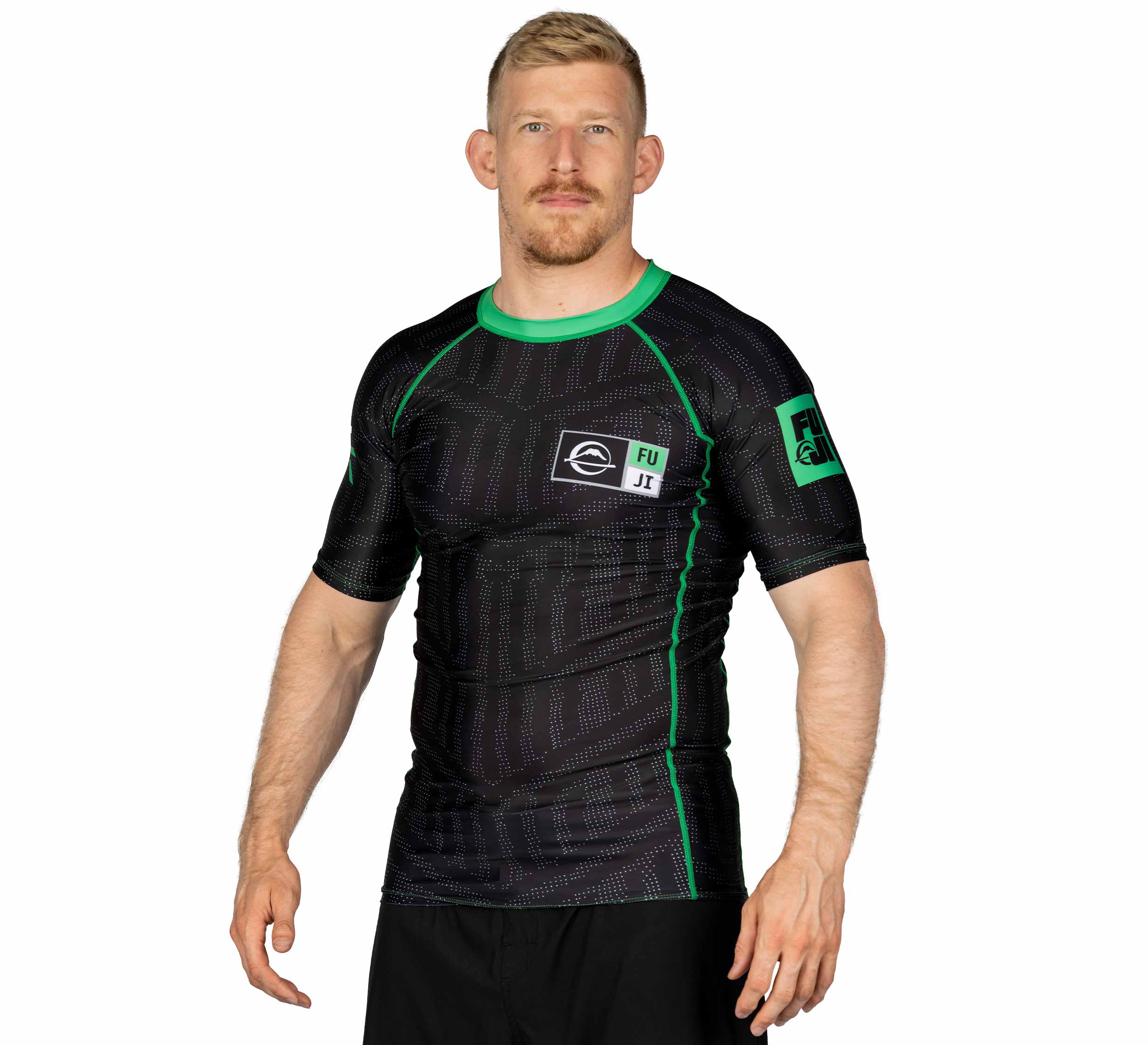 A man with short blond hair and a beard wears the Team FUJI Short Sleeve Rashguard Green and black shorts, standing against a white background.