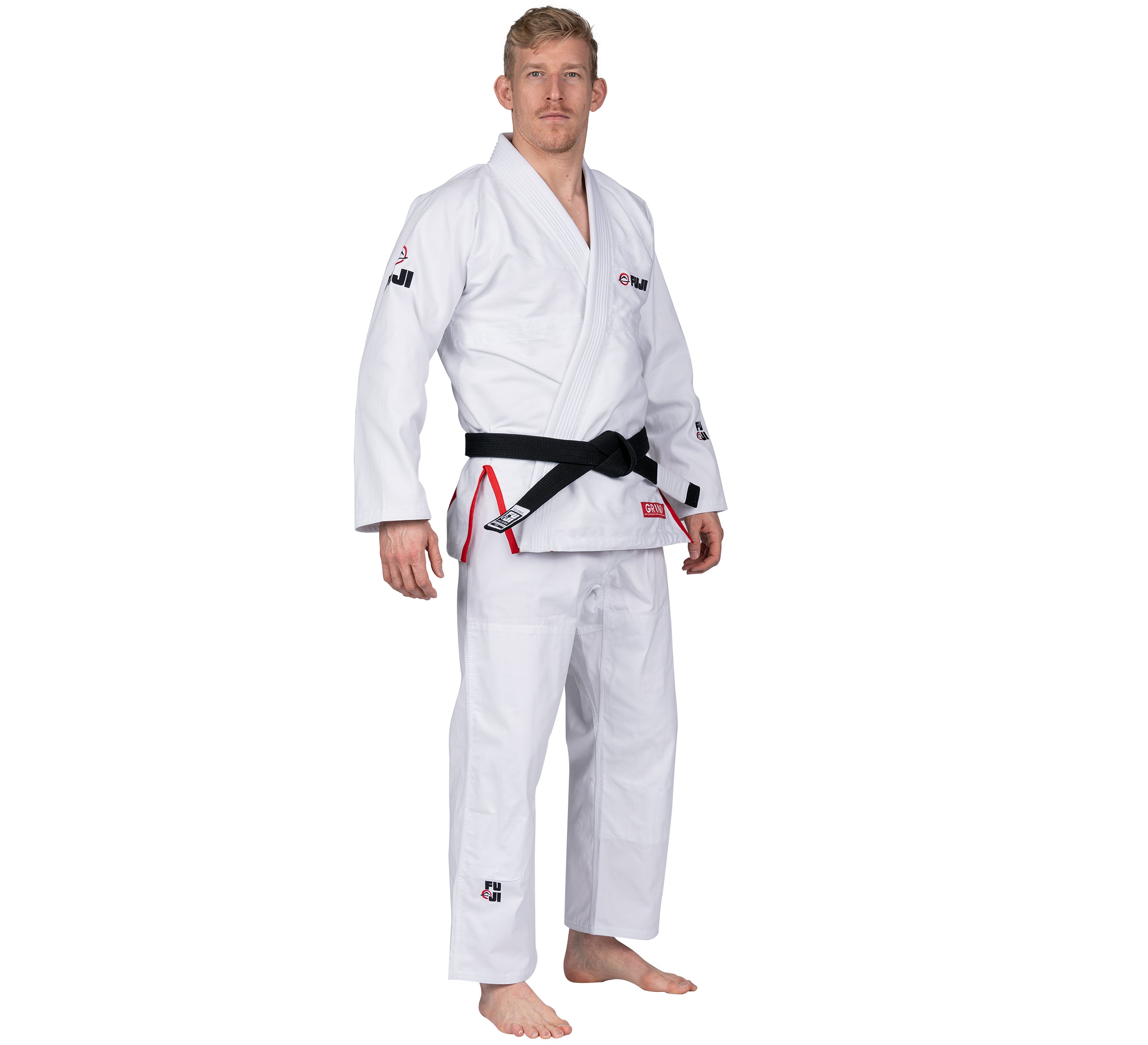 A man stands barefoot, wearing the Elemental BJJ Gi Grind White with a black belt, looking directly at the camera against a plain white background.