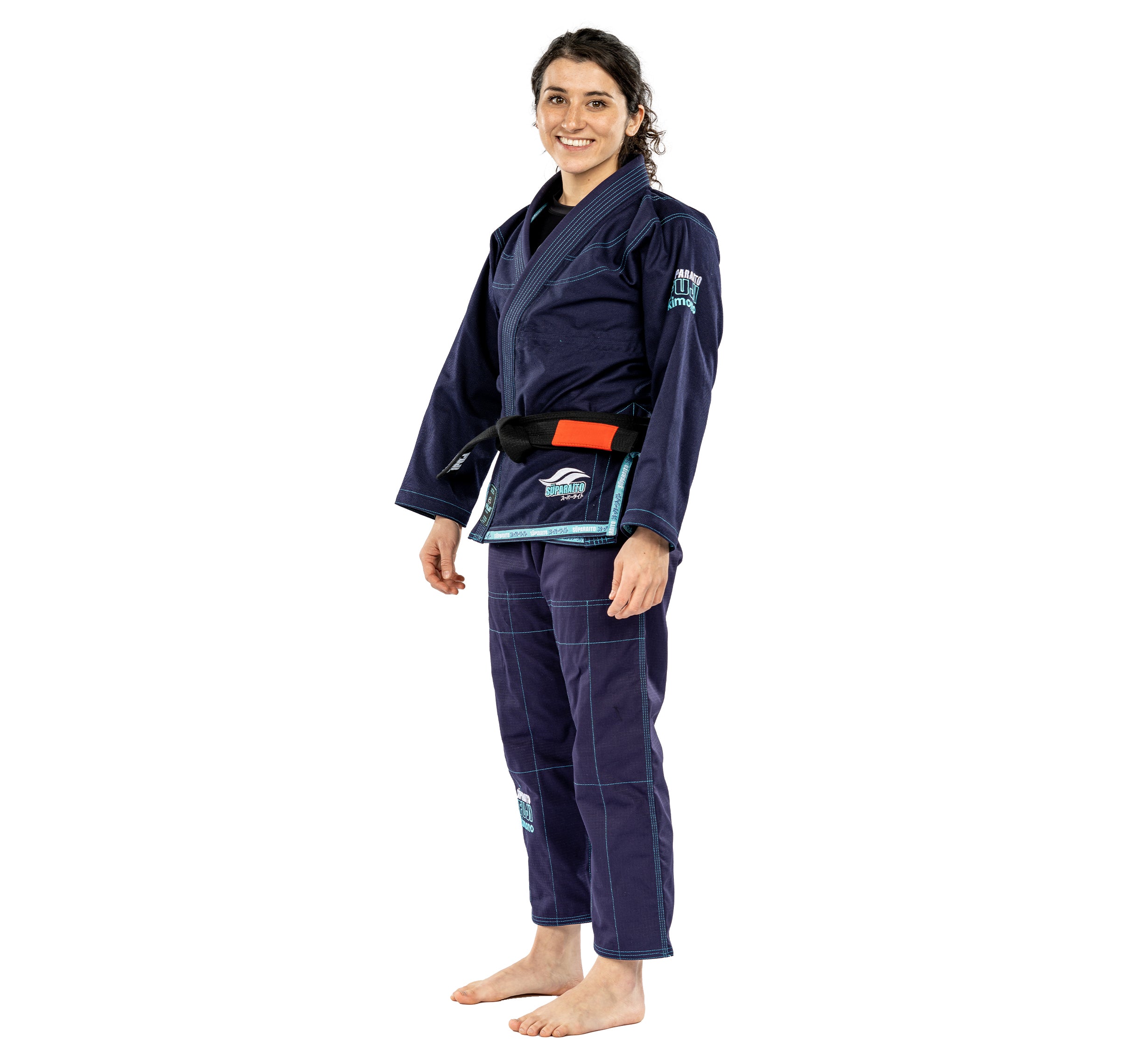 A smiling person stands barefoot against a white background, wearing the Suparaito Womens BJJ Gi Navy with a black belt.