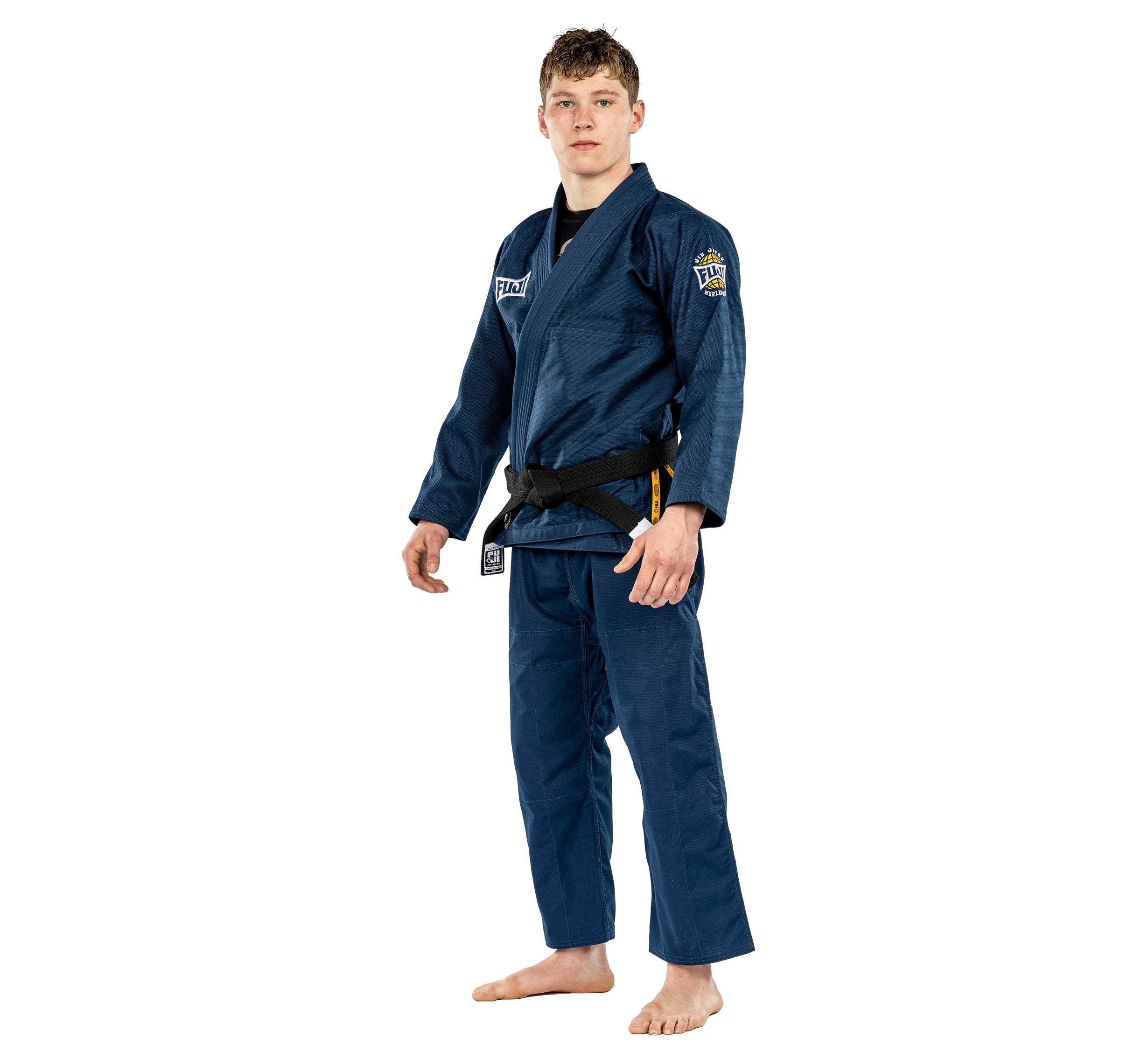 A young man stands barefoot on a white background, wearing the Suparaito BJJ Gi Worldwide Edition Navy with patches and a black belt, looking forward with a neutral expression.