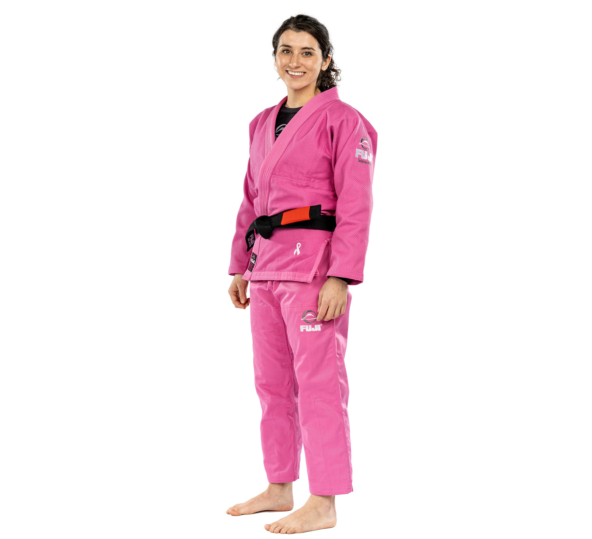 A person stands barefoot, smiling, and wearing the All Around Womens BJJ Gi Pink with a black belt. The gi features white logos and a ribbon symbol on the chest and shoulder, set against a plain white background.