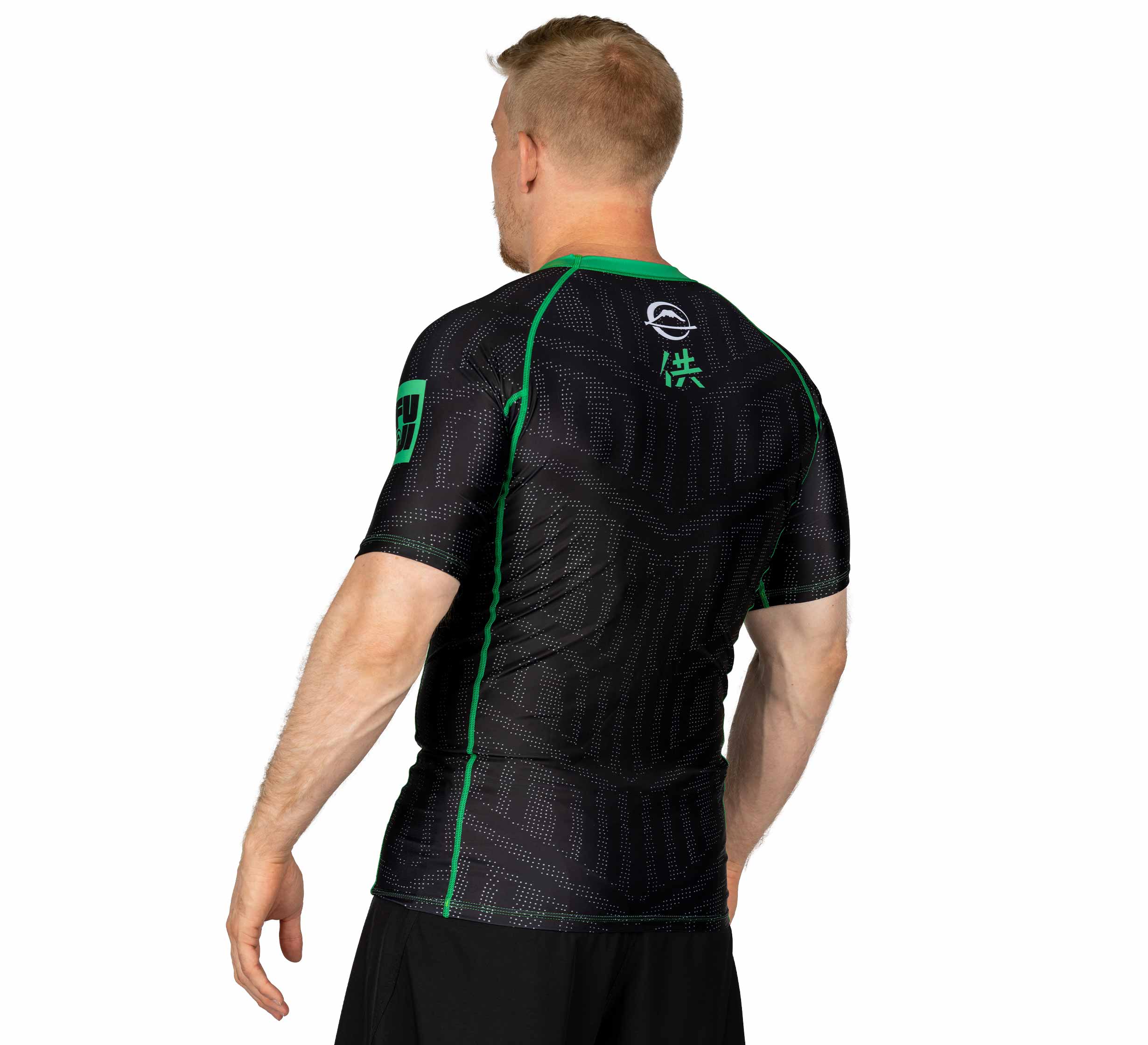 A man with short blond hair wears the Team FUJI Short Sleeve Rashguard Green, featuring black fabric and green seams, standing facing away with his left arm slightly extended against a plain white background.