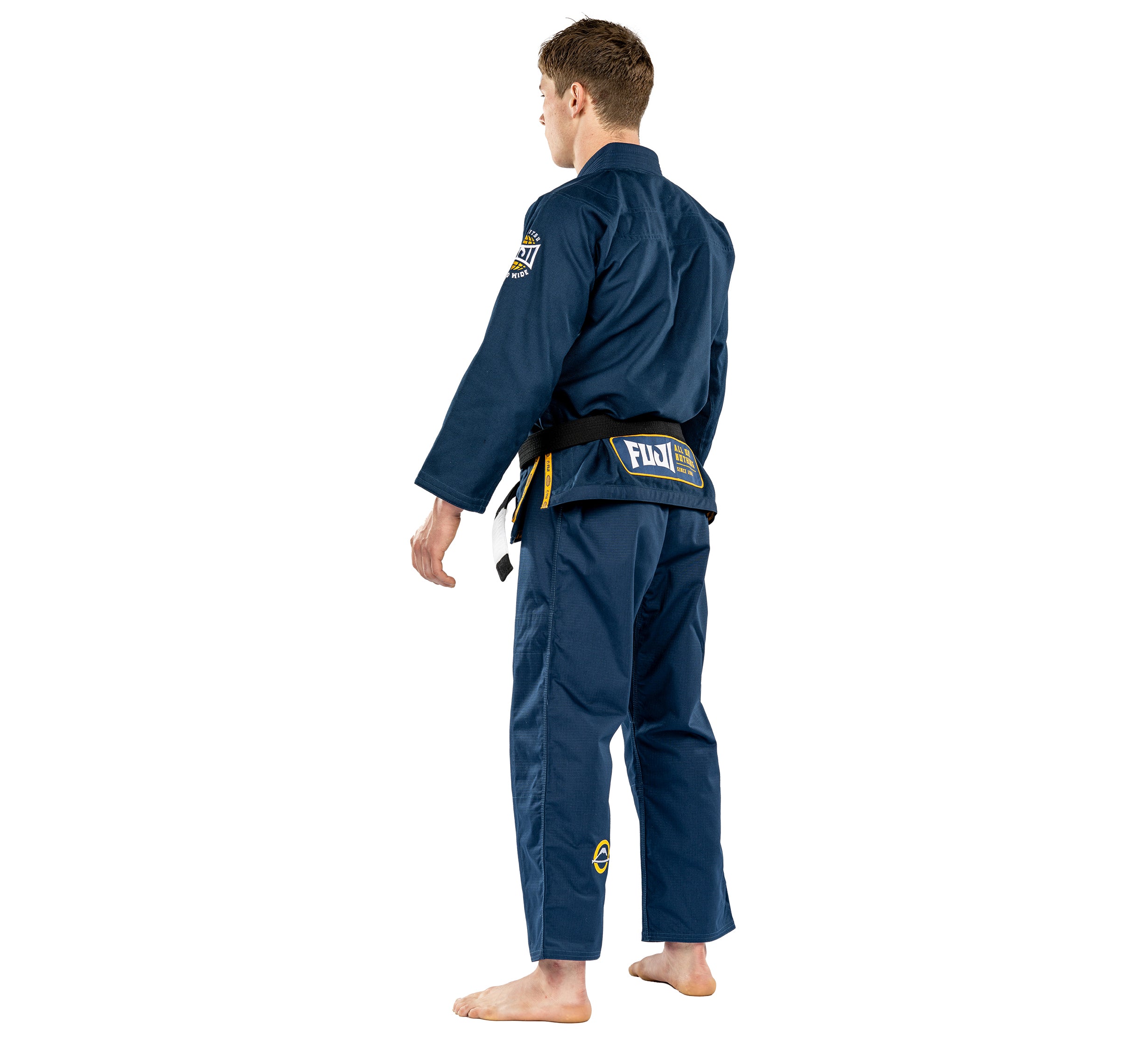 A man stands barefoot facing away, wearing the Suparaito BJJ Gi Worldwide Edition Navy with a black belt and patches, hands relaxed at his sides.