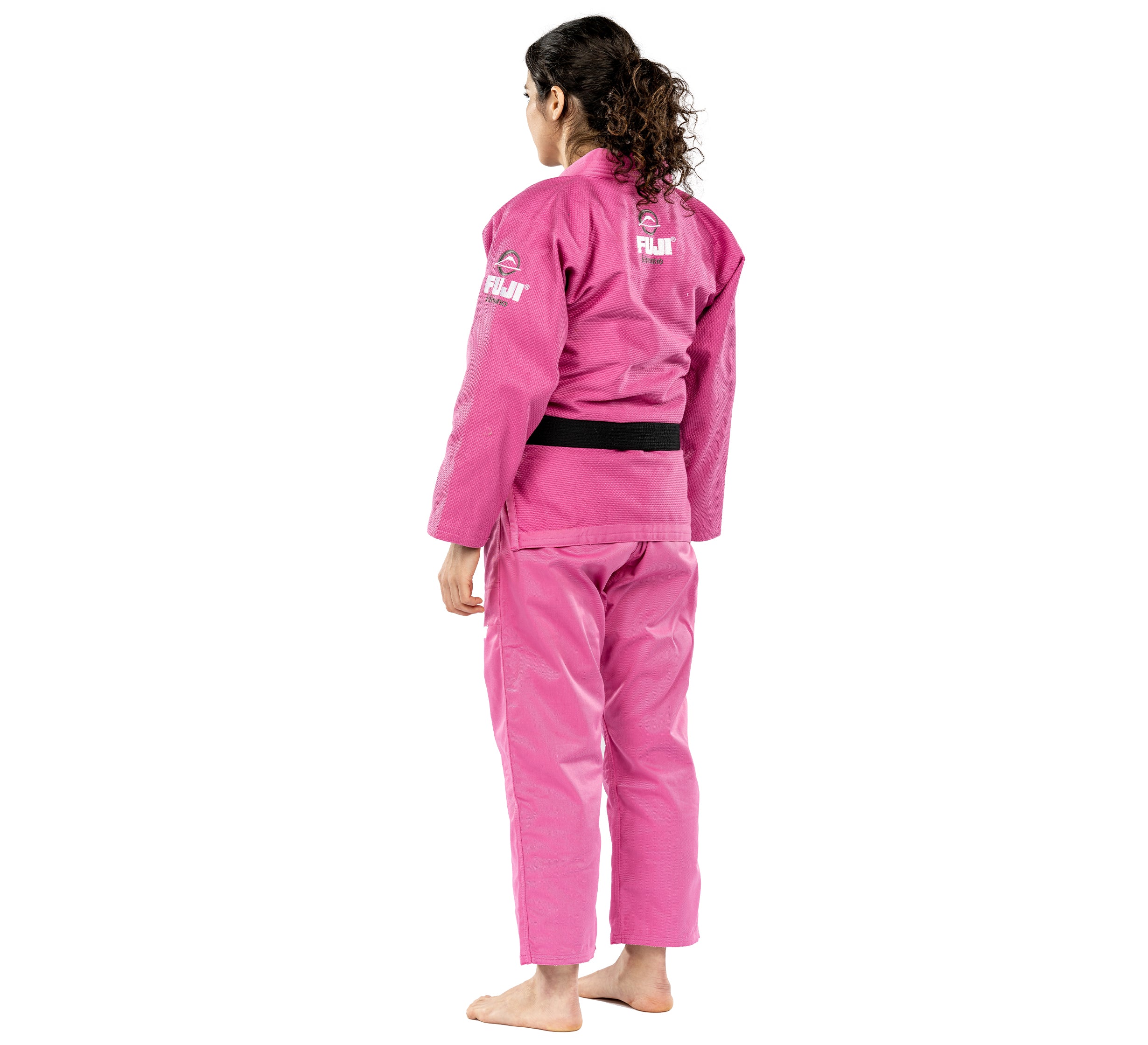 A person with long curly hair stands barefoot, facing away with head turned slightly, wearing the All Around Womens BJJ Gi Pink and a black belt.