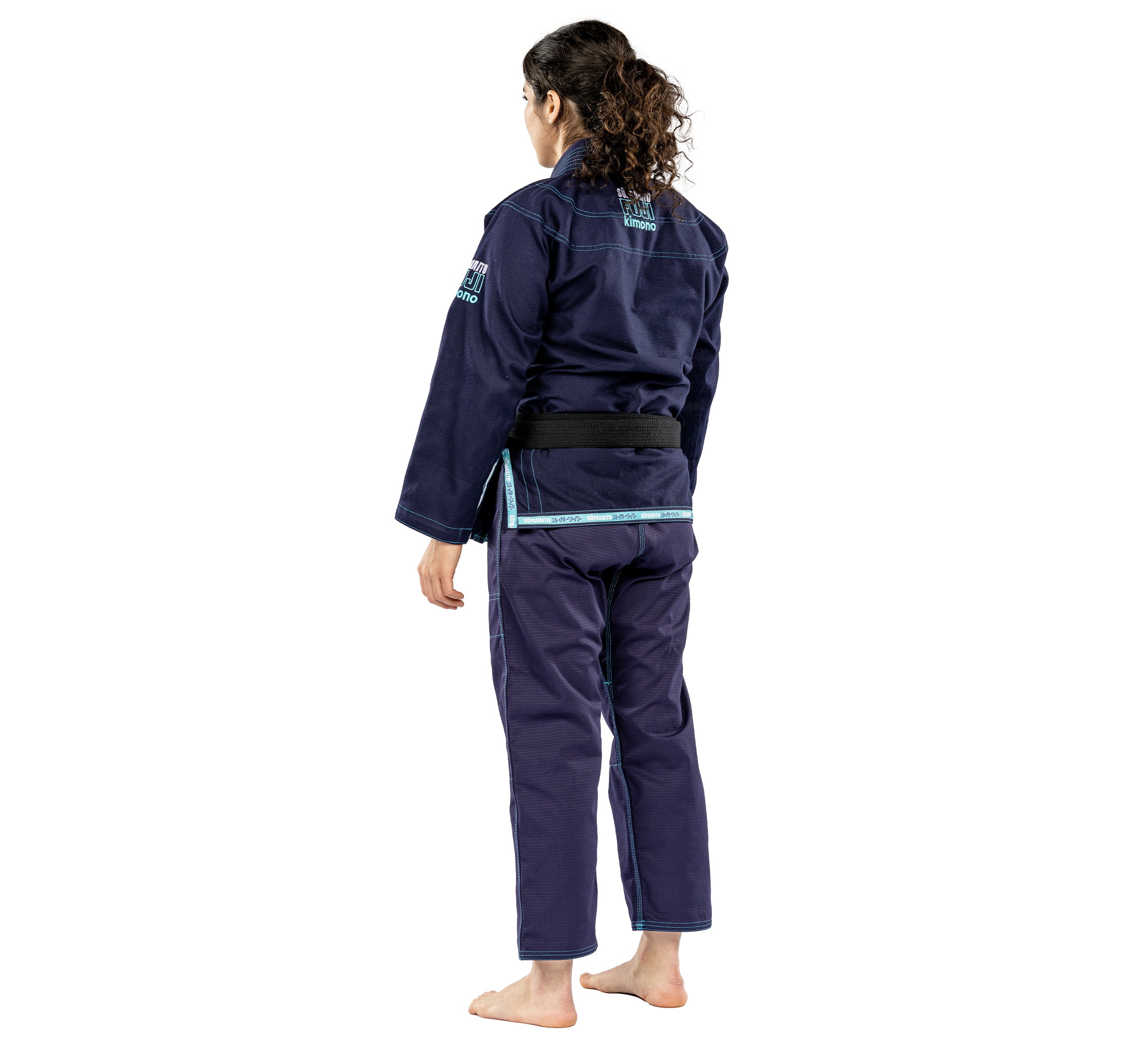A person with curly hair stands barefoot, facing away, wearing the Suparaito Womens BJJ Gi Navy and a black belt.