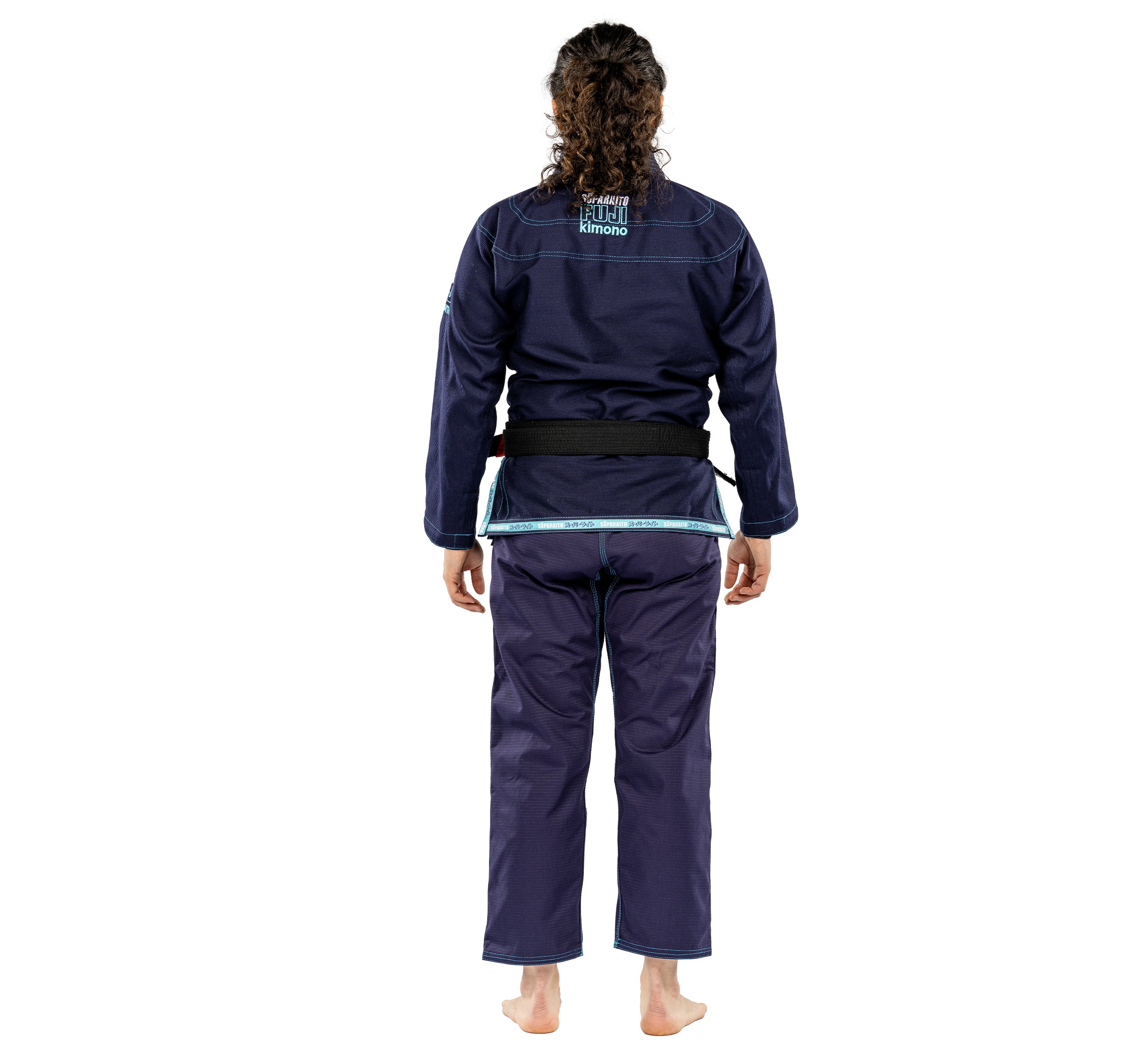 A person with curly hair stands barefoot, facing away from the camera in a Suparaito Womens BJJ Gi Navy and a black belt, set against a white background.