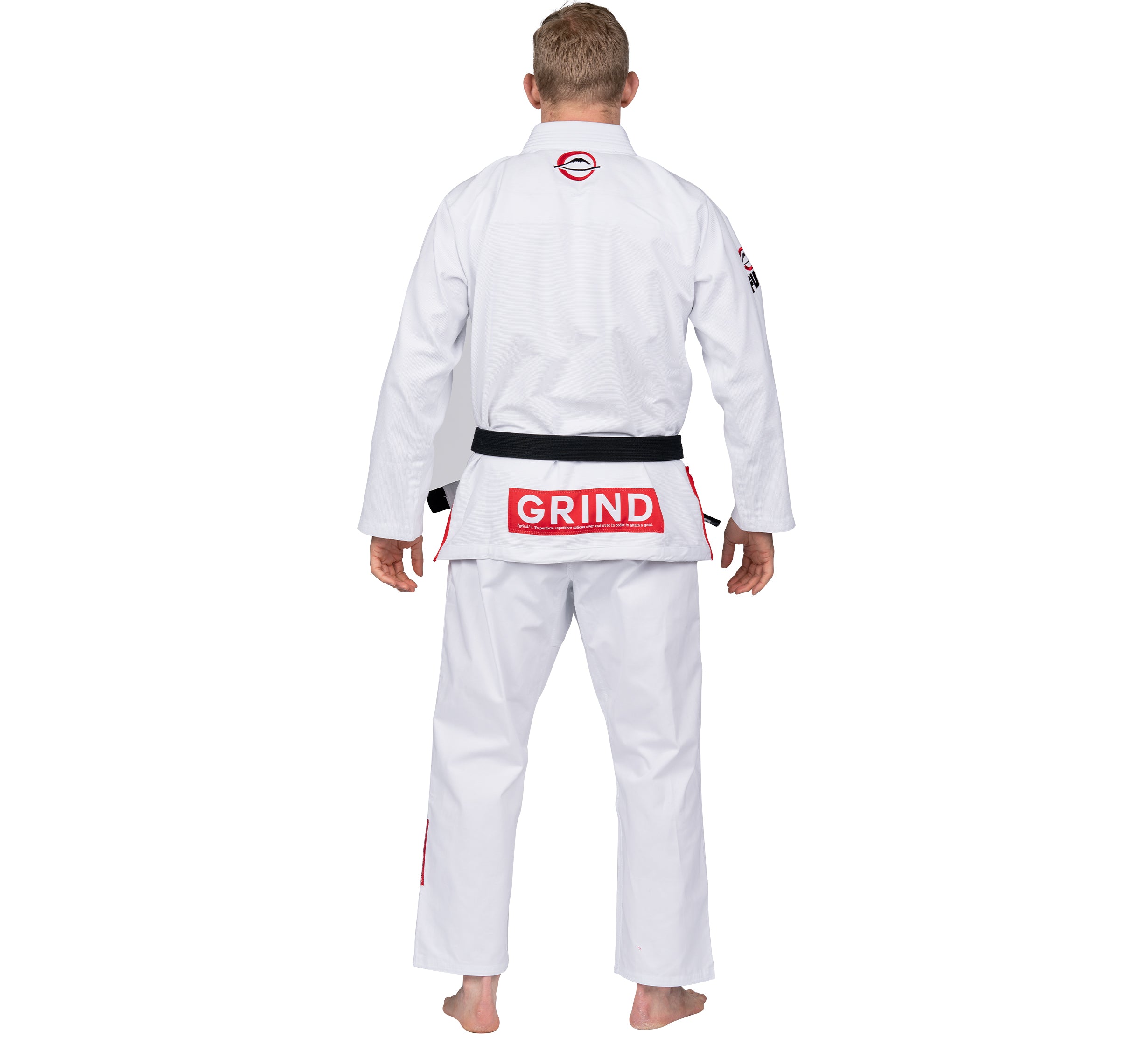 A person stands barefoot facing away in the Elemental BJJ Gi Grind White, a white martial arts uniform with black belt, featuring a red GRIND patch and circular logo on the upper back.