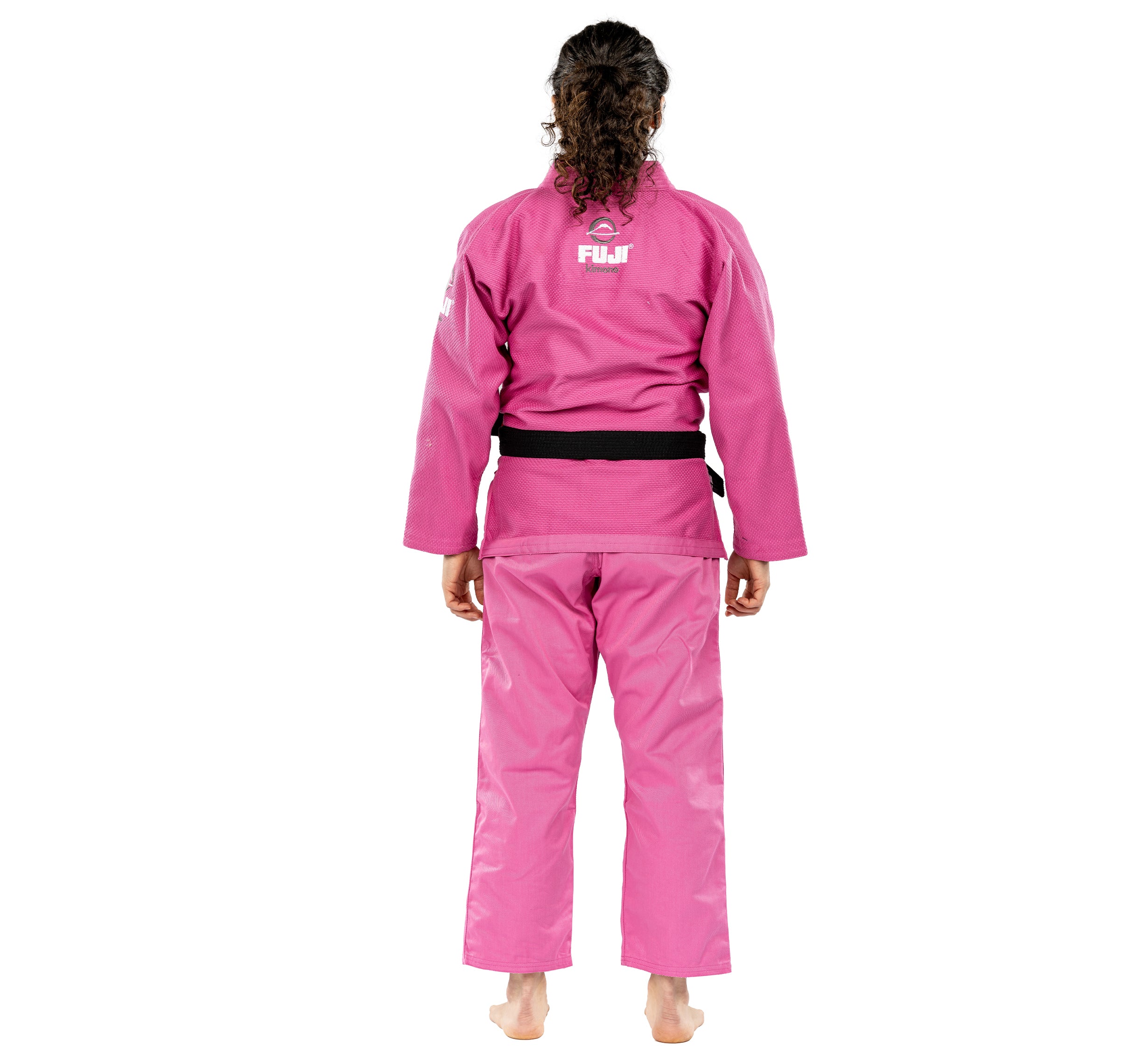 A person with curly hair stands barefoot on a plain white background, facing away and wearing the All Around Womens BJJ Gi Pink with a black belt. "FUJI" is written on the back of the gi.