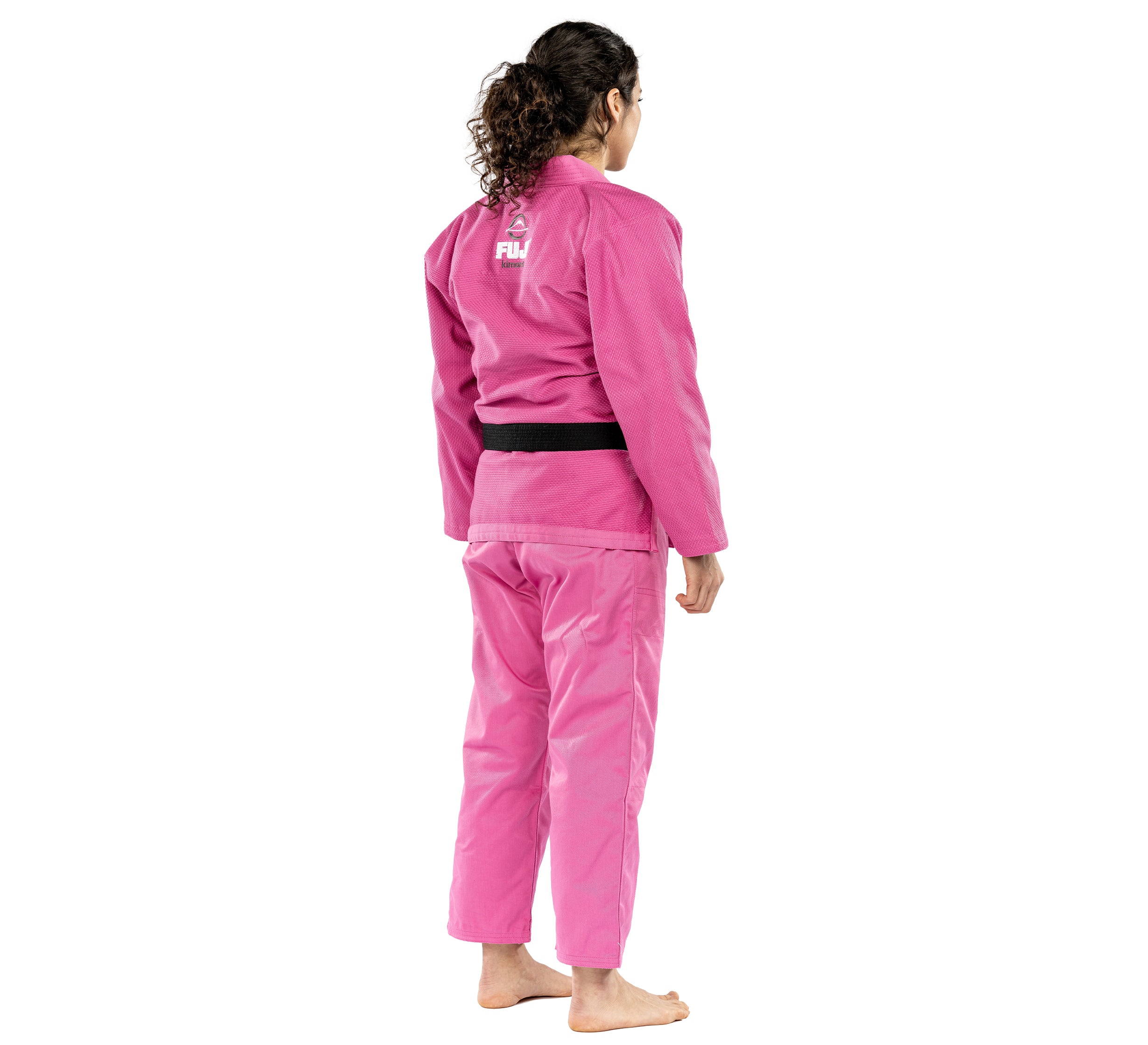 A person with curly hair, barefoot and wearing the All Around Womens BJJ Gi Pink with a black belt, stands facing away against a white background.